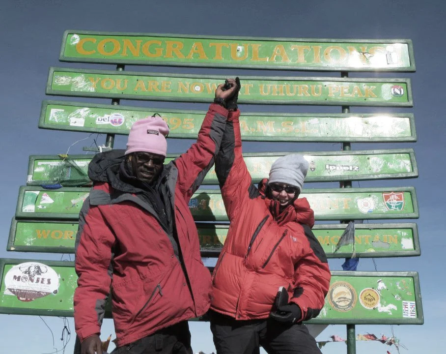 Two climbers wearing red jackets and gray hats celebrate at Uhuru Peak, Mount Kilimanjaro, with a green sign behind them reading 'Congratulations! You are now at Uhuru Peak, 5895 m.'