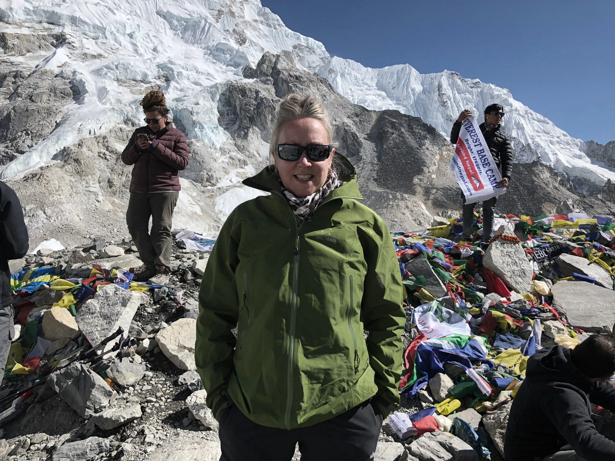 A woman in a green jacket and sunglasses taking a photo on a mountain summit. Behind her are other climbers, one holding a flag and another looking at their phone, with snowy mountains and rocks around.