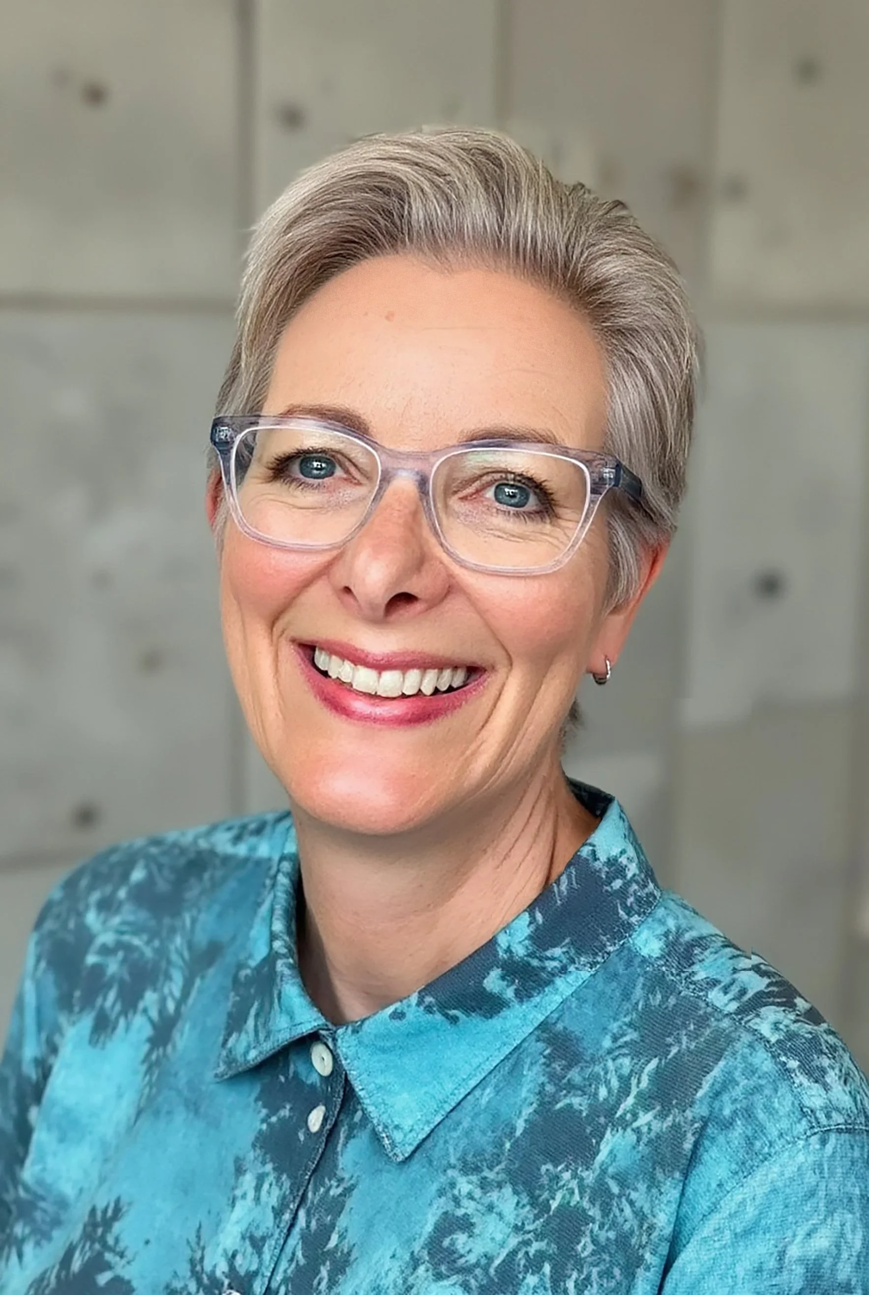 A portrait of a smiling woman with short gray hair, wearing clear glasses and a blue patterned shirt, in front of a blurred gray background.