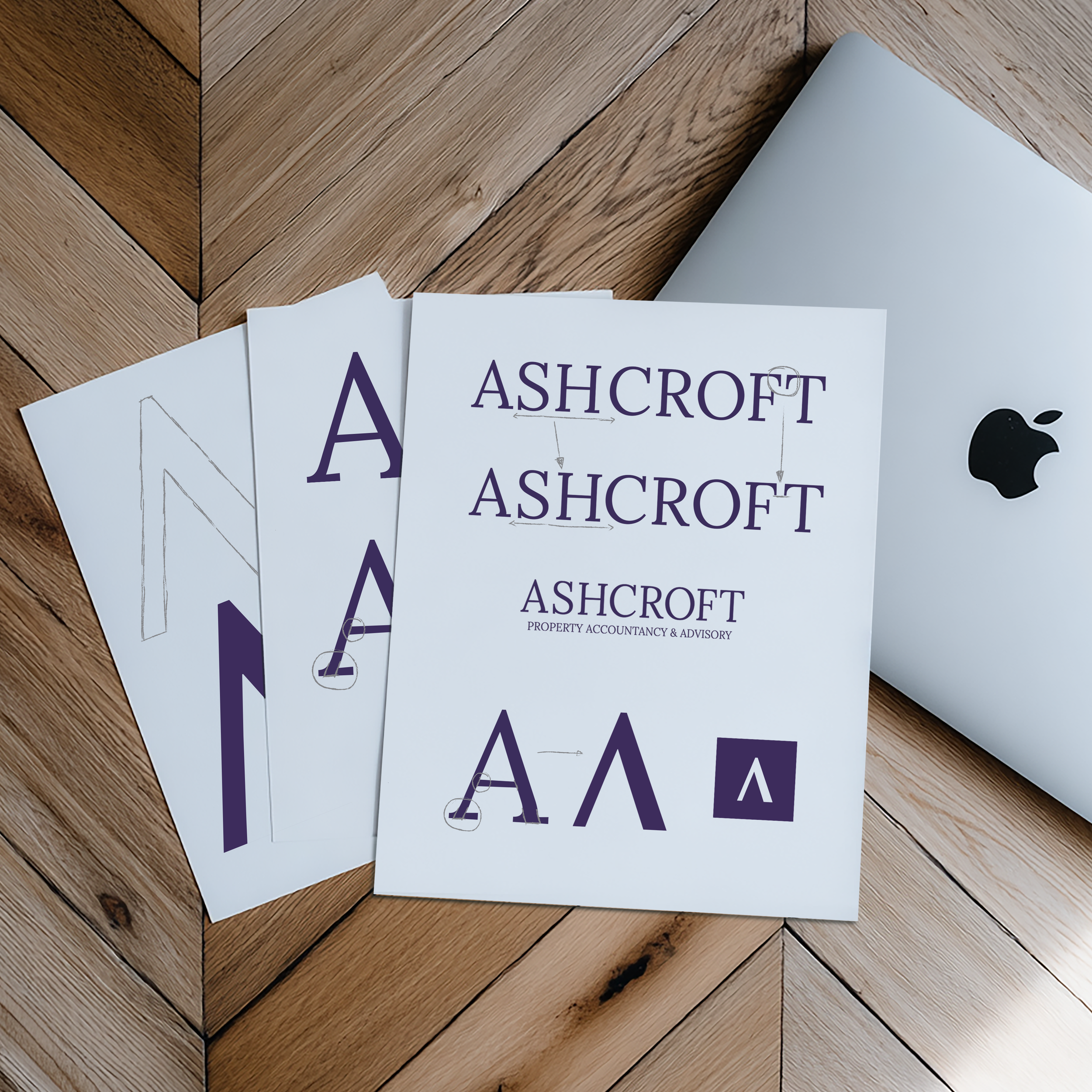 Printed sheets with the logo and branding for Ashcroft Property Accountancy & Advisory, featuring the company's name and stylized 'A' logo, placed on a wooden surface alongside a closed silver laptop with an Apple logo.
