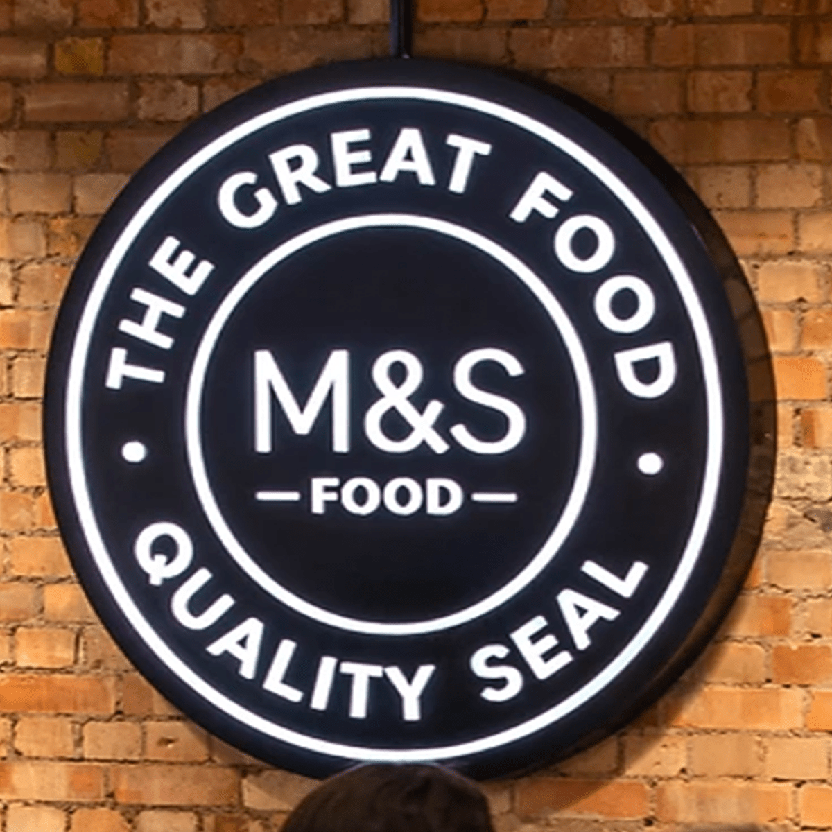 Illuminated circular sign with the text 'The Great Food QALITY SEAL' and 'M&S Food' in the center, mounted on a brick wall.