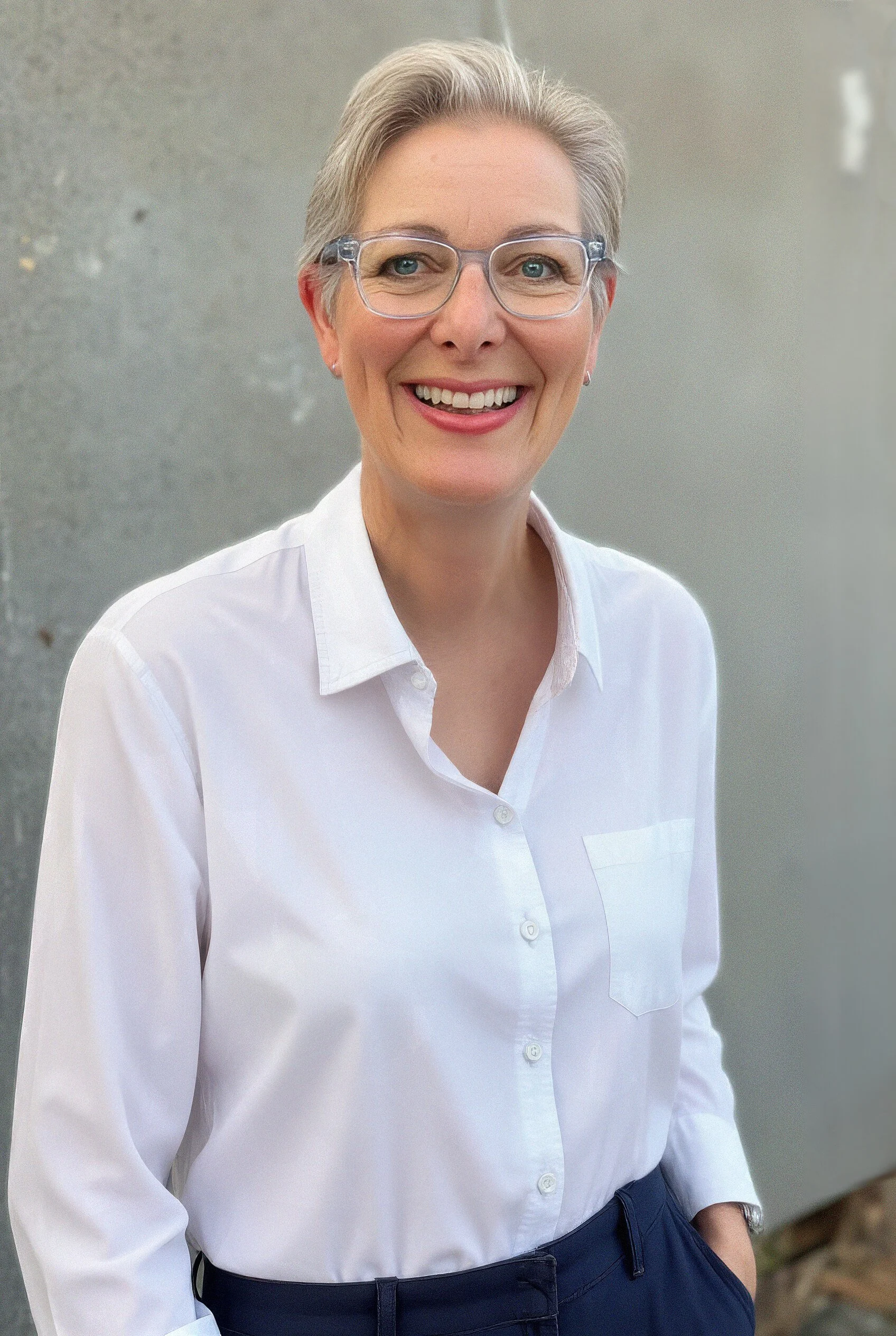 A smiling woman with short gray hair and glasses wearing a white button-up shirt and dark pants standing outdoors.