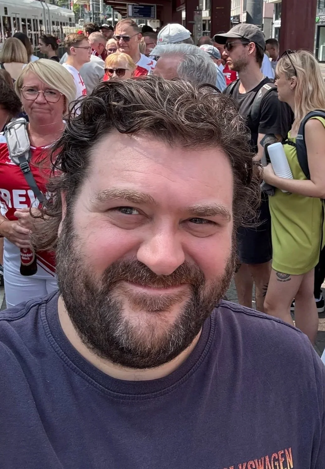 A man with brown curly hair and a beard taking a selfie in a crowded outdoor area with many people around him, some wearing backpacks and casual clothing.