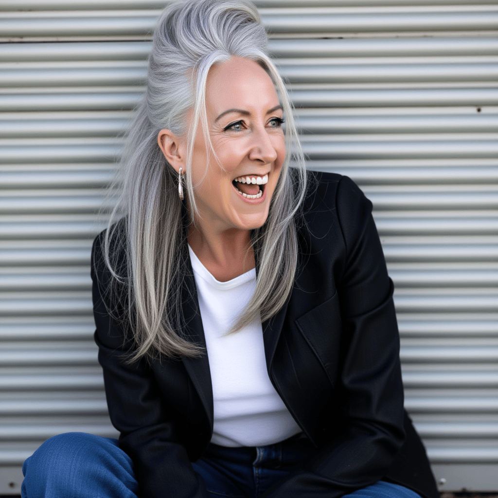 A woman with long gray hair laughing outdoors against a metal background, wearing a black blazer and white top.
