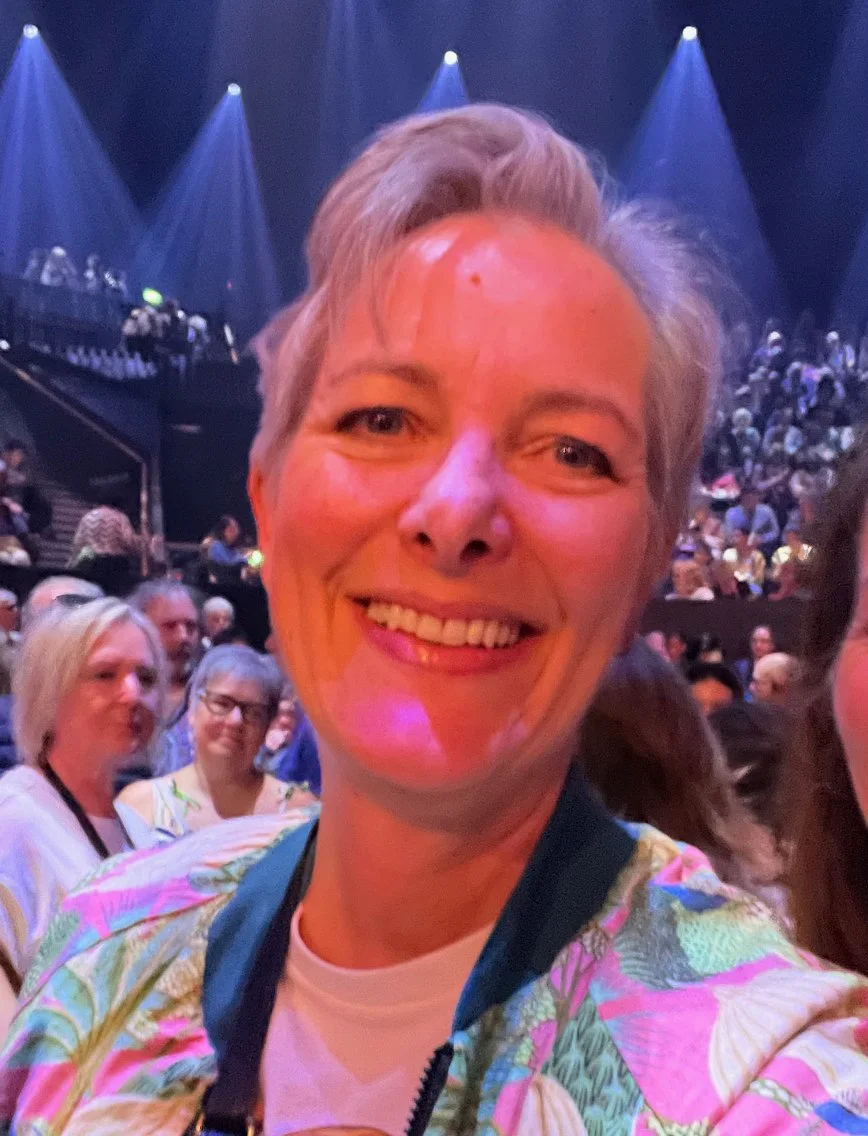 A smiling woman taking a selfie at a crowded indoor event or concert with a stage and spotlights in the background.