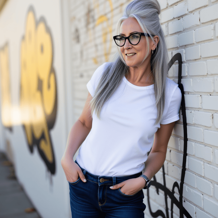 A silver-haired woman with glasses, wearing a white t-shirt and jeans, leaning against a white brick wall with graffiti, smiling outdoors.