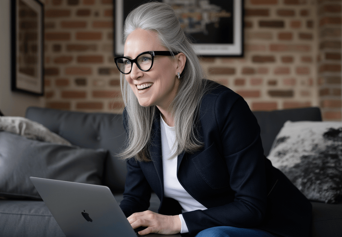 A woman with gray hair, glasses, and earrings smiling while using a laptop in a living room with a brick wall and framed pictures.