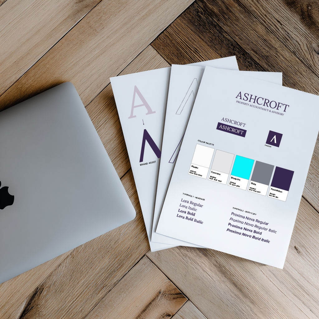 Three printed branding and design guides for Ashcroft, a property accounting and advisory firm, are spread out on a wooden desk next to a closed MacBook laptop.