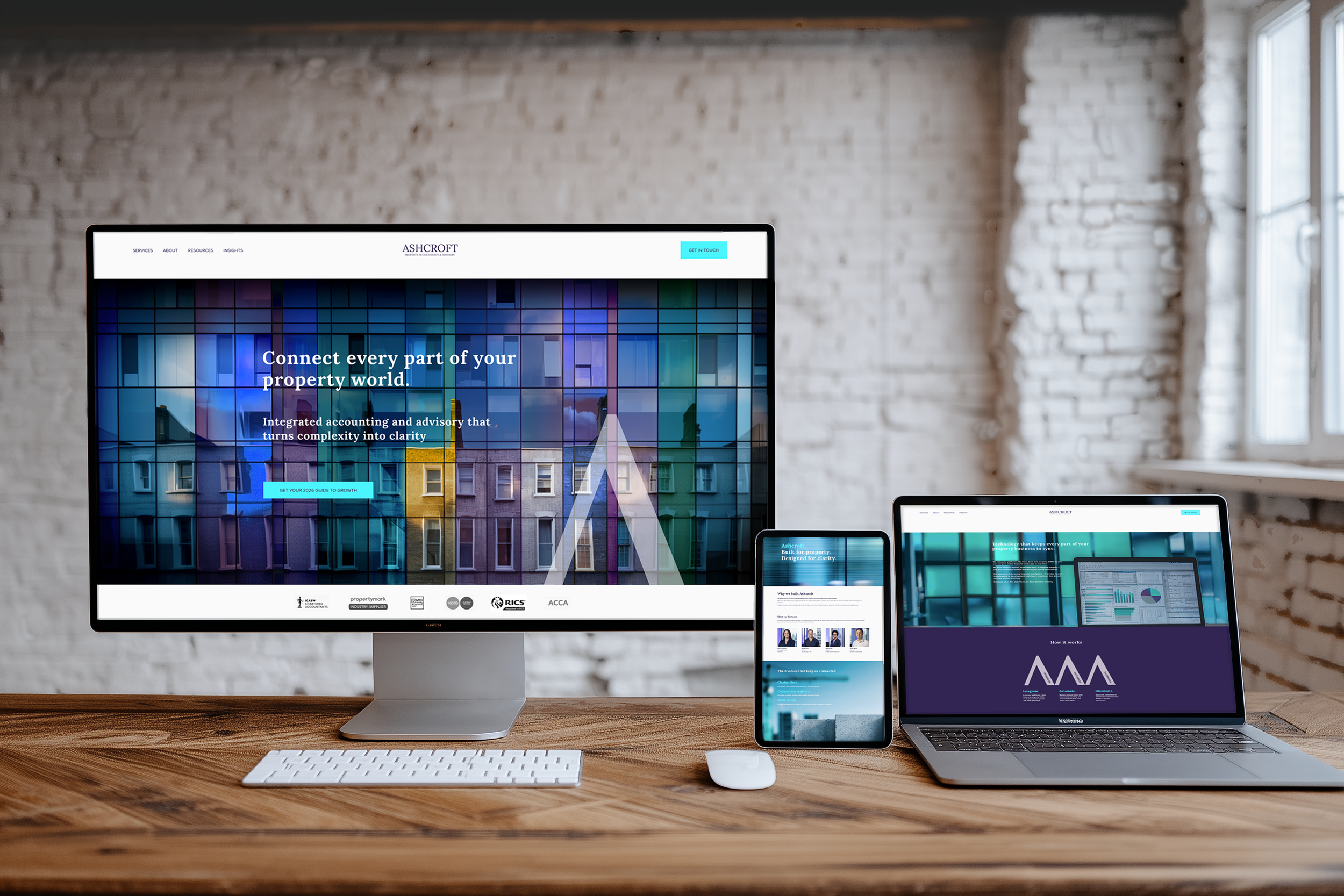 A workstation with a desktop computer, a laptop, and a smartphone on a wooden desk against a brick wall. The desktop screen displays a colorful website with the text 'Connect every part of your property world.' The laptop shows a website with a bar graph and pie chart, and the smartphone displays a website with profile pictures and text.