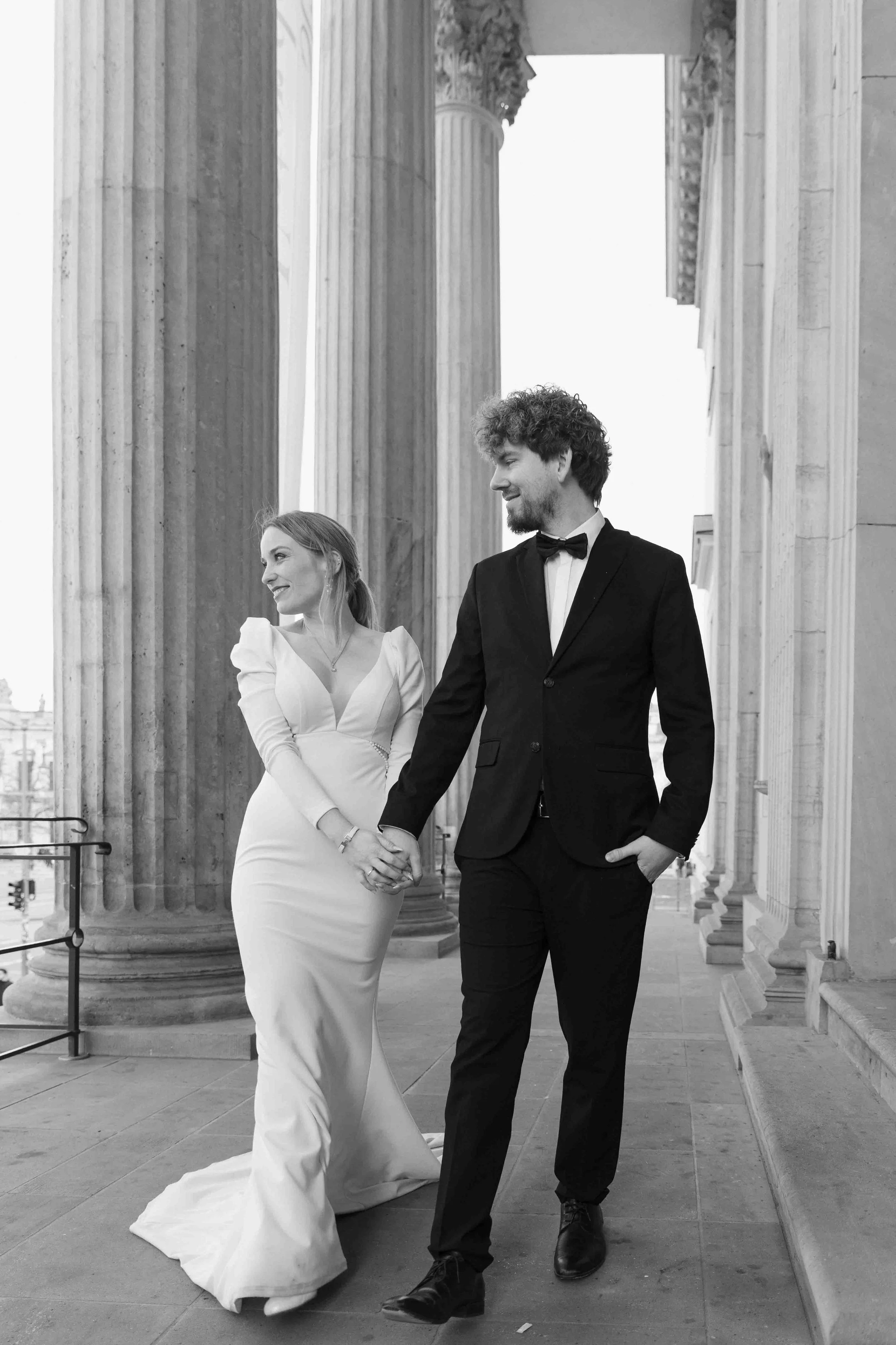 A couple dressed in wedding attire walking hand in hand outside a classical building with large columns, smiling at each other.