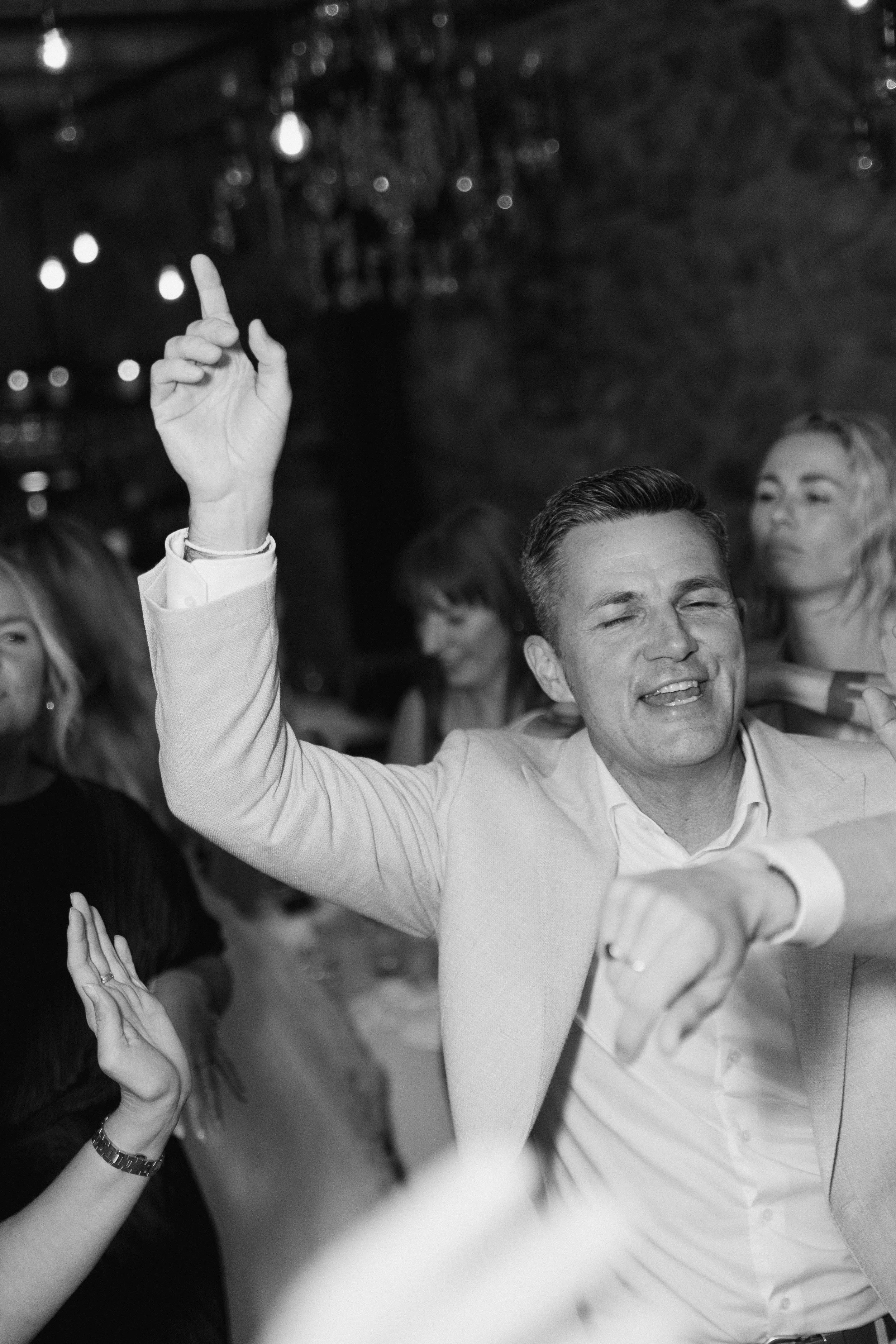 A man in a light-colored suit with his eyes closed, smiling, with his right arm raised and his index finger pointing up at a social gathering. There are blurred people and string lights in the background.