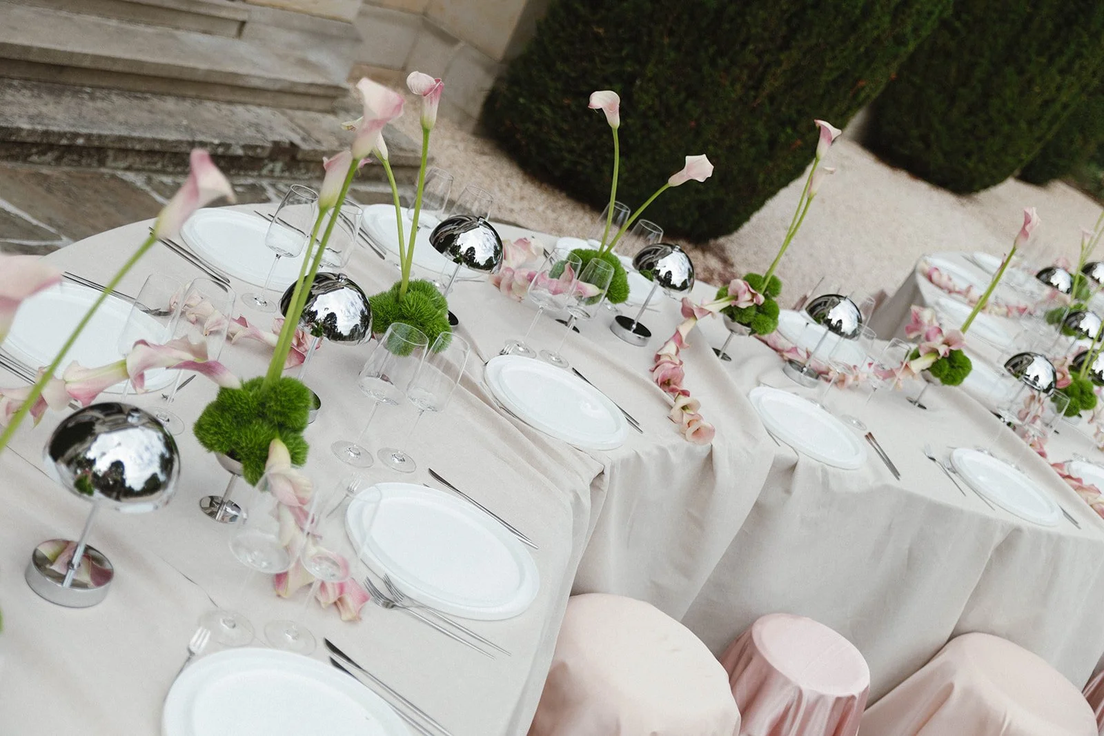 A long dining table set for an outdoor event, decorated with pink and white flowers, greenery, and shiny silver ornaments, with white plates and glassware arranged neatly.