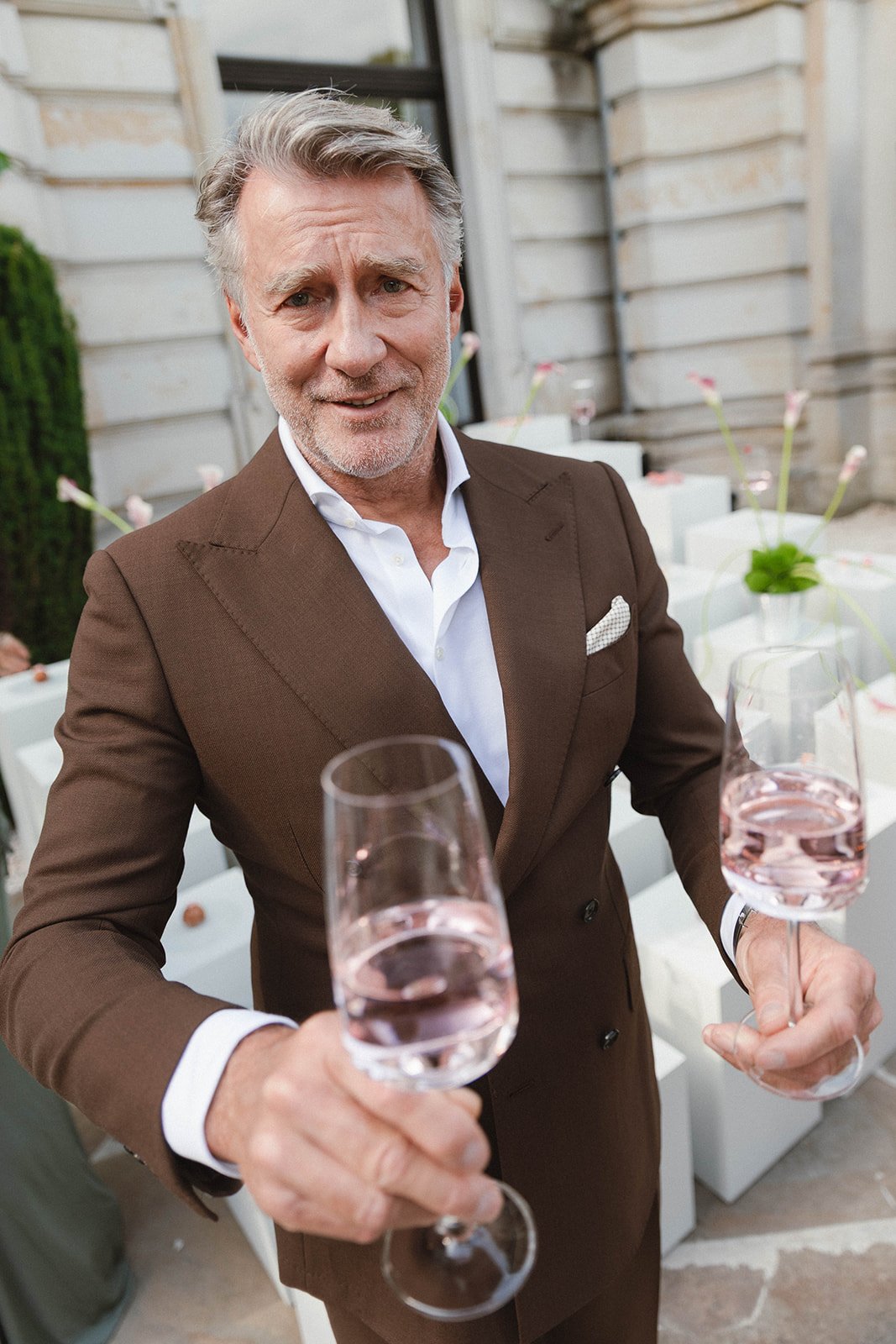 Older man in a brown suit holding two glasses of rosé wine during a wedding celebration or social event. Documentary and loose editorial style wedding photography.
