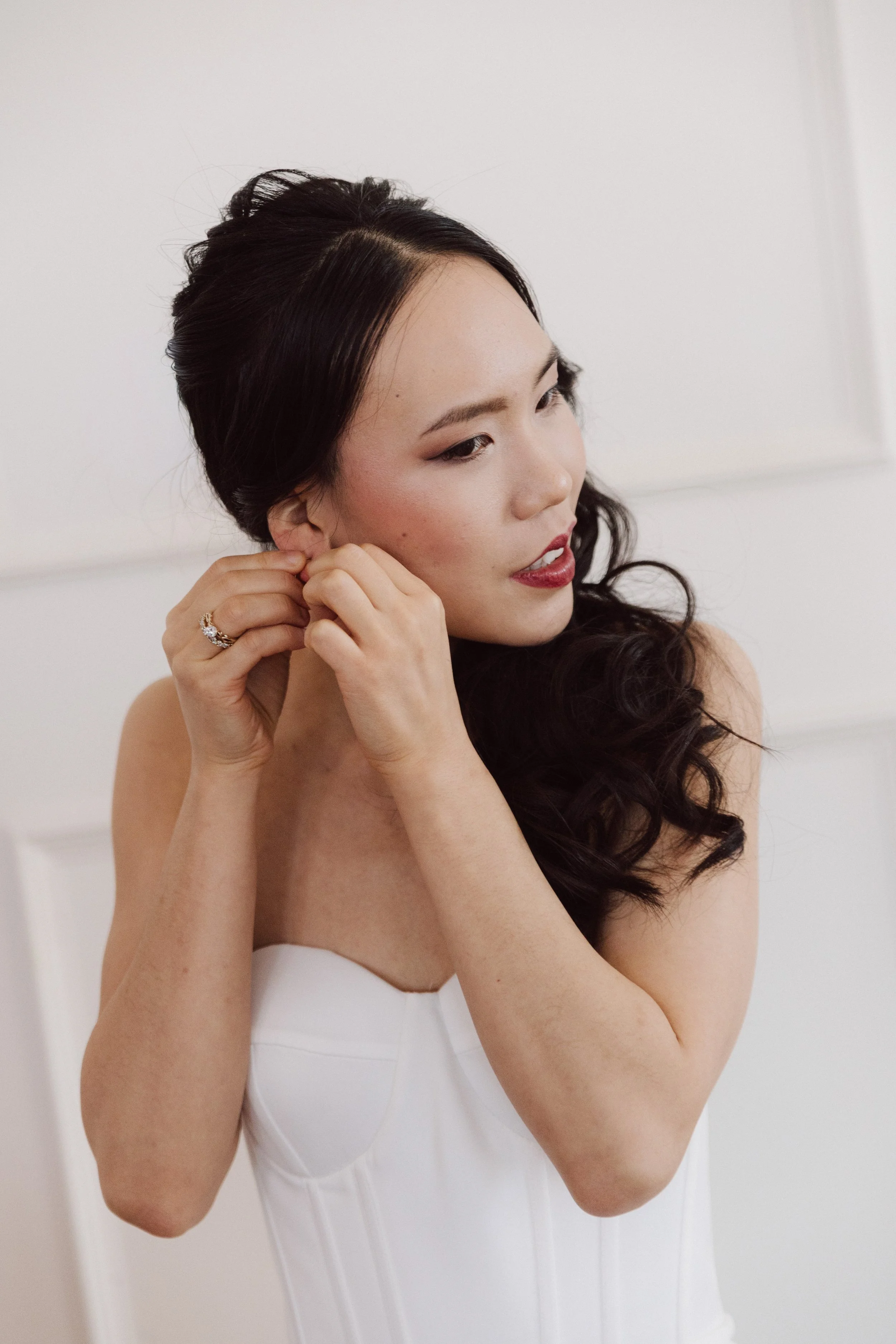Black haired bride putting in her jewellery before her wedding in a clean white gown before a clean white wall.