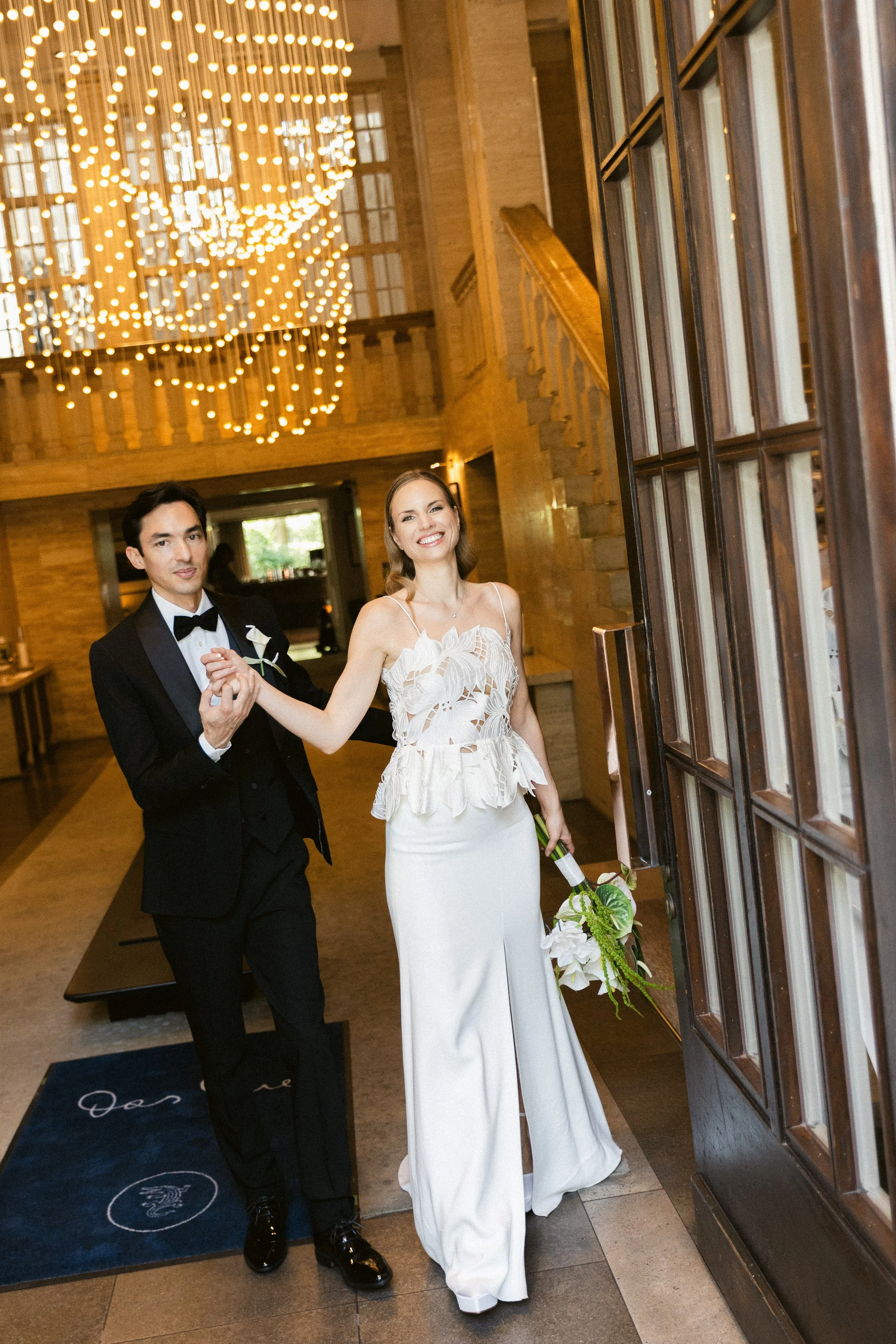 A newlywed couple, a man in a black tuxedo and a woman in a white wedding dress, is holding hands and entering a venue with warm lighting, a chandelier, and wooden decor.