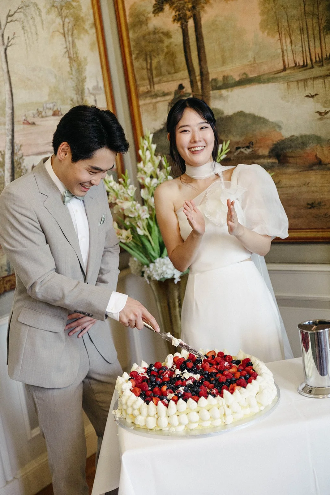 A man and woman are cutting a wedding cake with berries, while the woman claps and smiles in an elegant room with floral arrangements and landscape paintings in the background. Documentary and loose editorial style wedding photography.