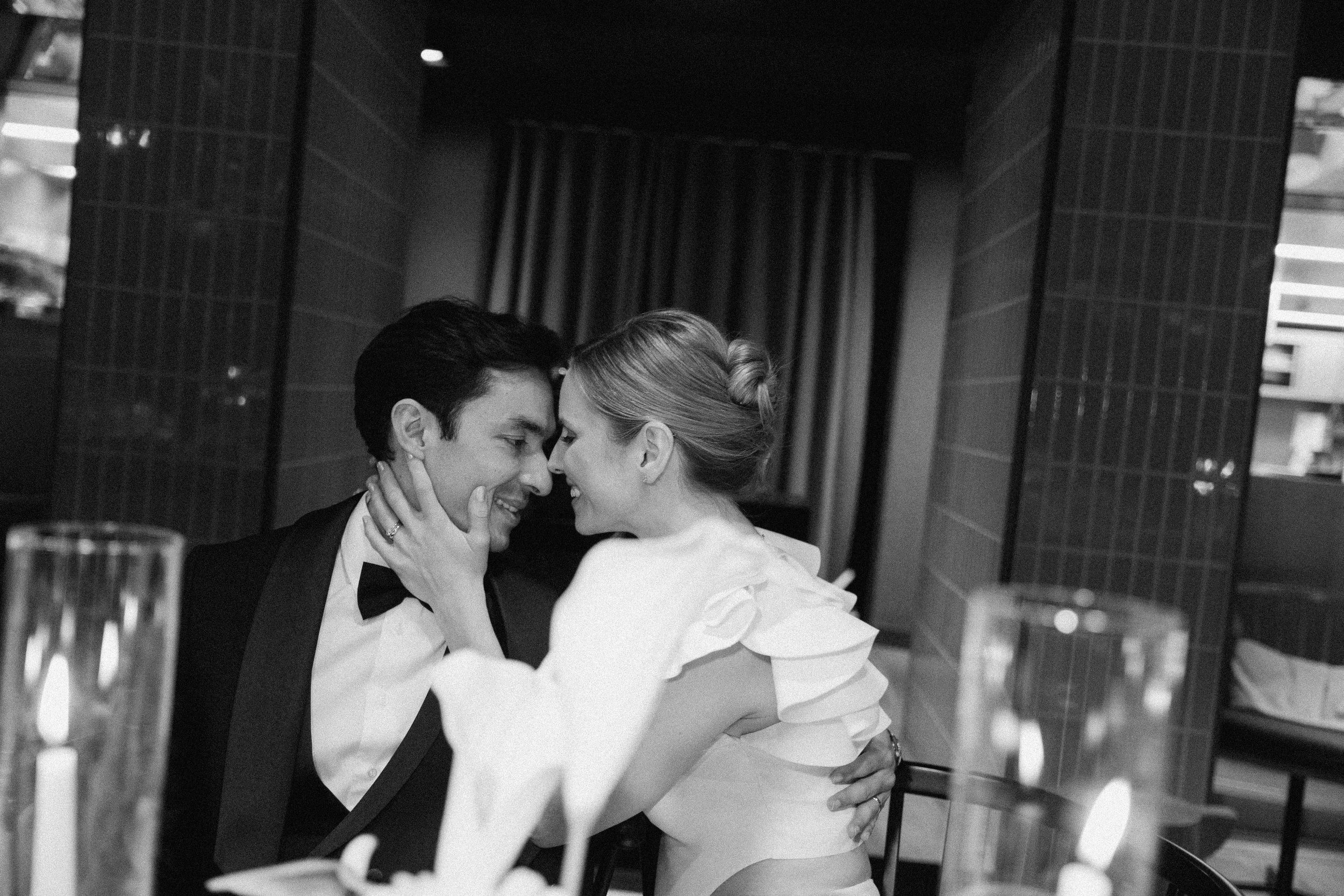 A black and white photo of a recently married couple sharing an intimate moment, touching foreheads, in a modern indoor setting with tiled walls and curtains in the background.