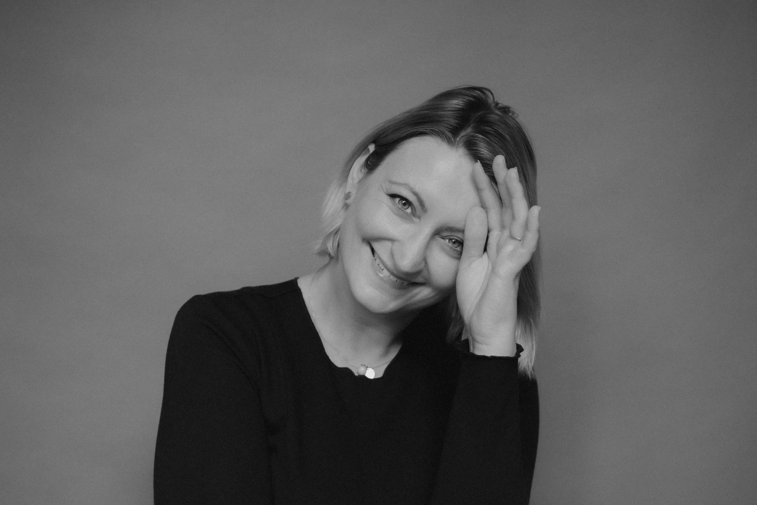 Black and white portrait of a smiling woman with short hair, her hand partially covering her face, against a plain background.