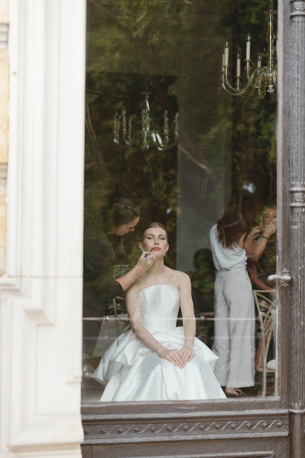 A woman in a white wedding dress is sitting in front of a mirror as her makeup is being applied by a makeup artist, with another woman adjusting her hair in the background, seen through a window or reflection. Documentary and loose editorial style we