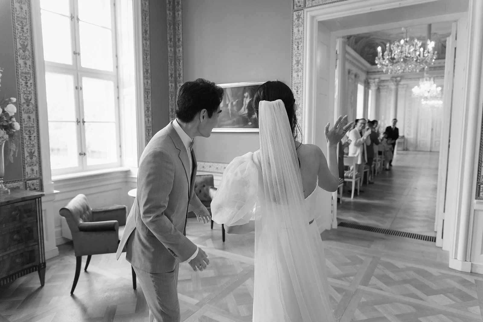 Black and white photo of a bride with a long veil and a groom in a suit, standing in a decorated, elegant room, with a group of people in the background. Documentary and loose editorial style wedding photography.