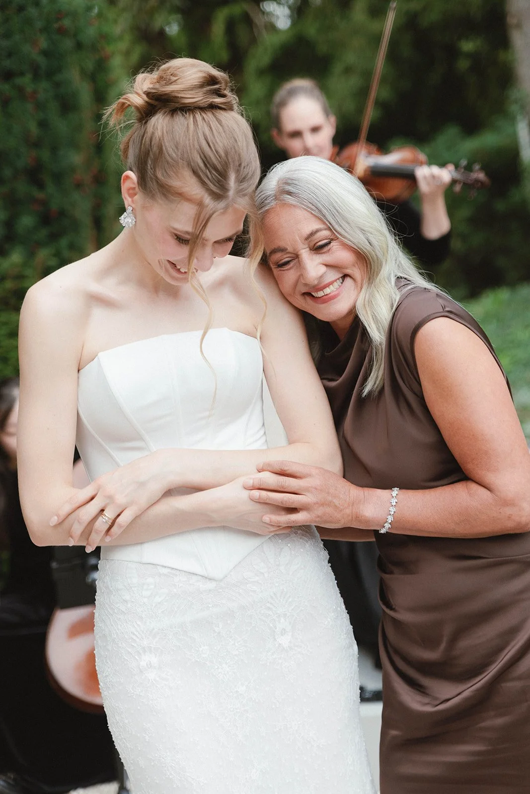 A young woman in a white wedding dress is smiling and looking down as she is held by an older woman with gray hair, wearing a brown dress, who is also smiling. A violinist is visible in the background playing outdoors. Documentary and loose editorial