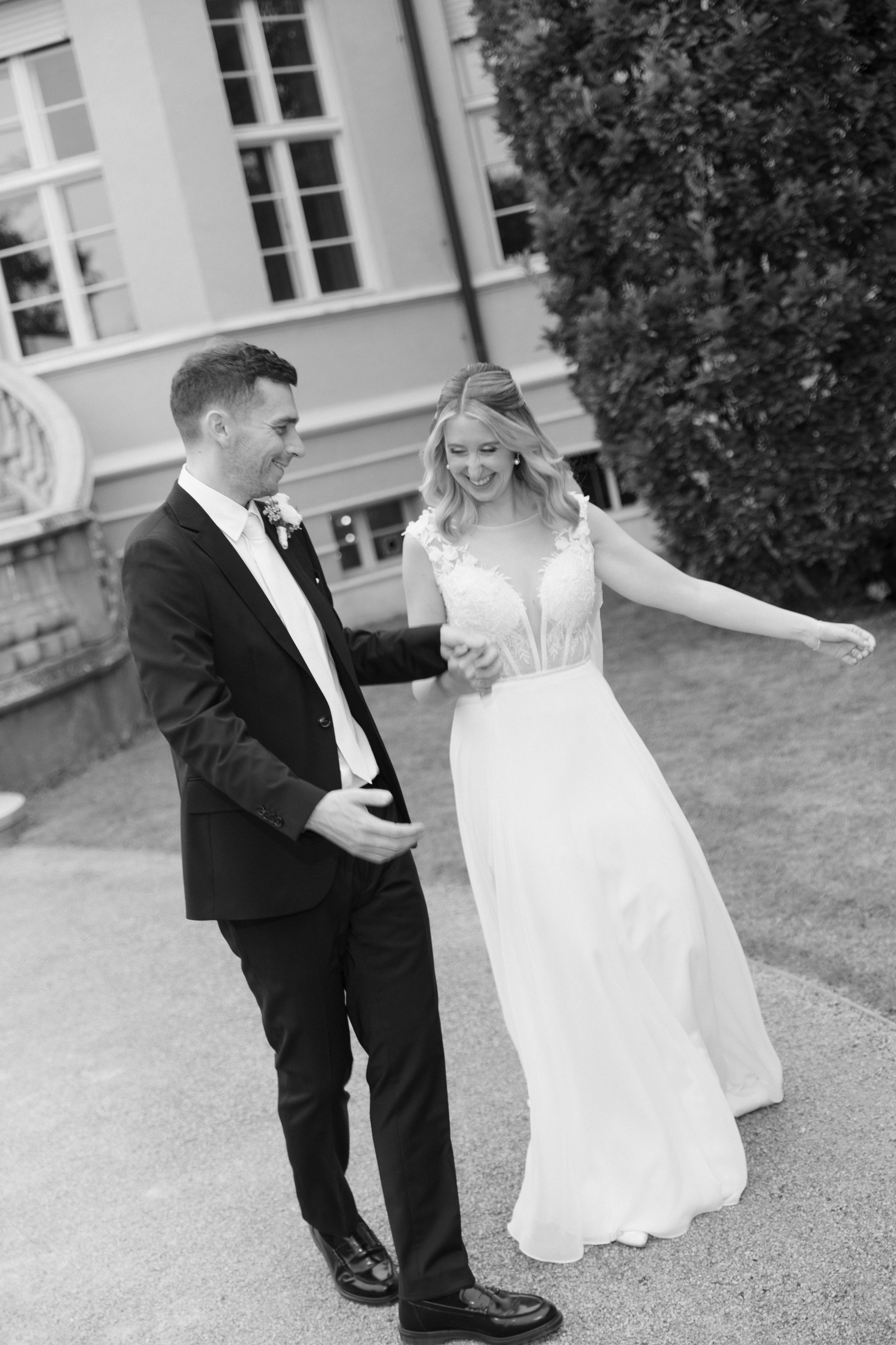 A bride and groom dancing outdoors in front of a building with large windows and a large bush. The groom is wearing a black suit, white shirt, and is smiling. The bride is in a white wedding dress with lace details and has blonde hair styled in loose