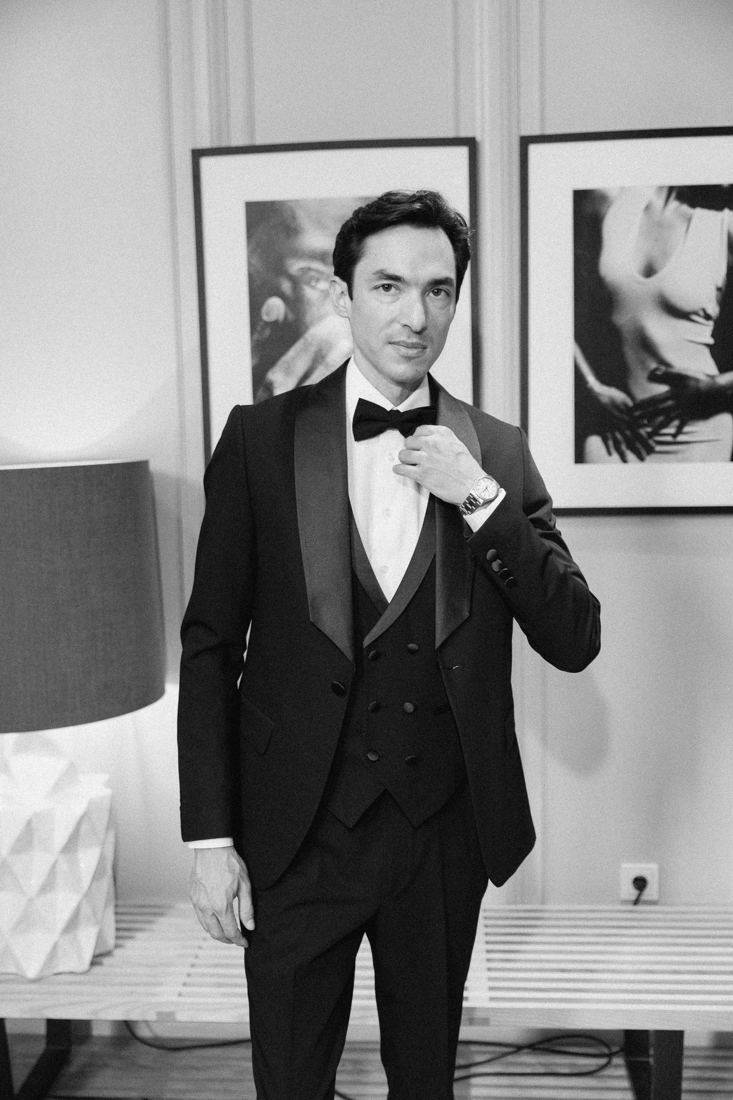 A man wearing a tuxedo with a bow tie, adjusting his cufflink, standing in an art gallery with framed photographs on the wall behind him.