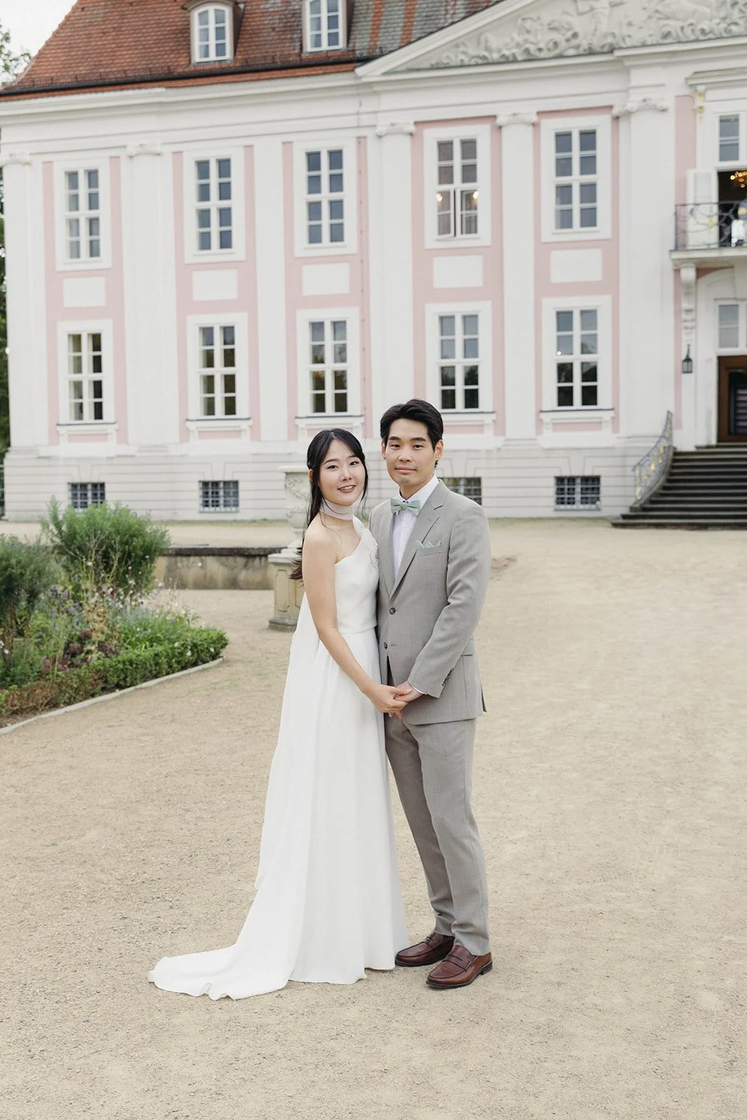 A bride and groom holding hands in front of a large pink and white historic building, with bushes and a gravel pathway.