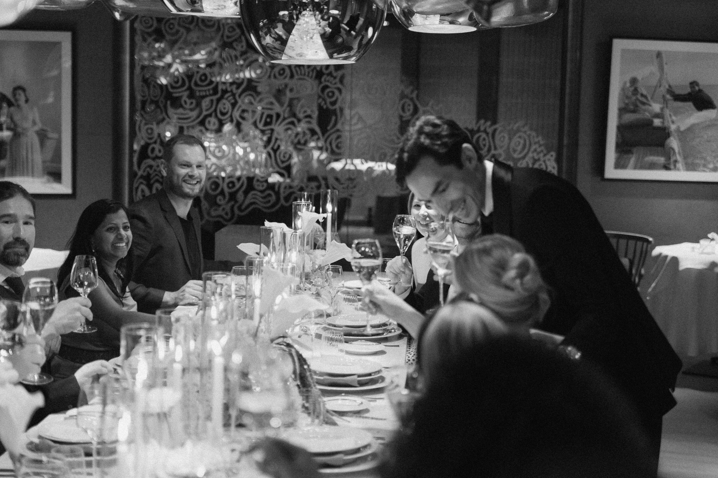 People of a wedding party sit at a long dining table with glasses and plates, enjoying a meal at a restaurant. A man in a suit is pouring a drink and others are smiling and conversing.