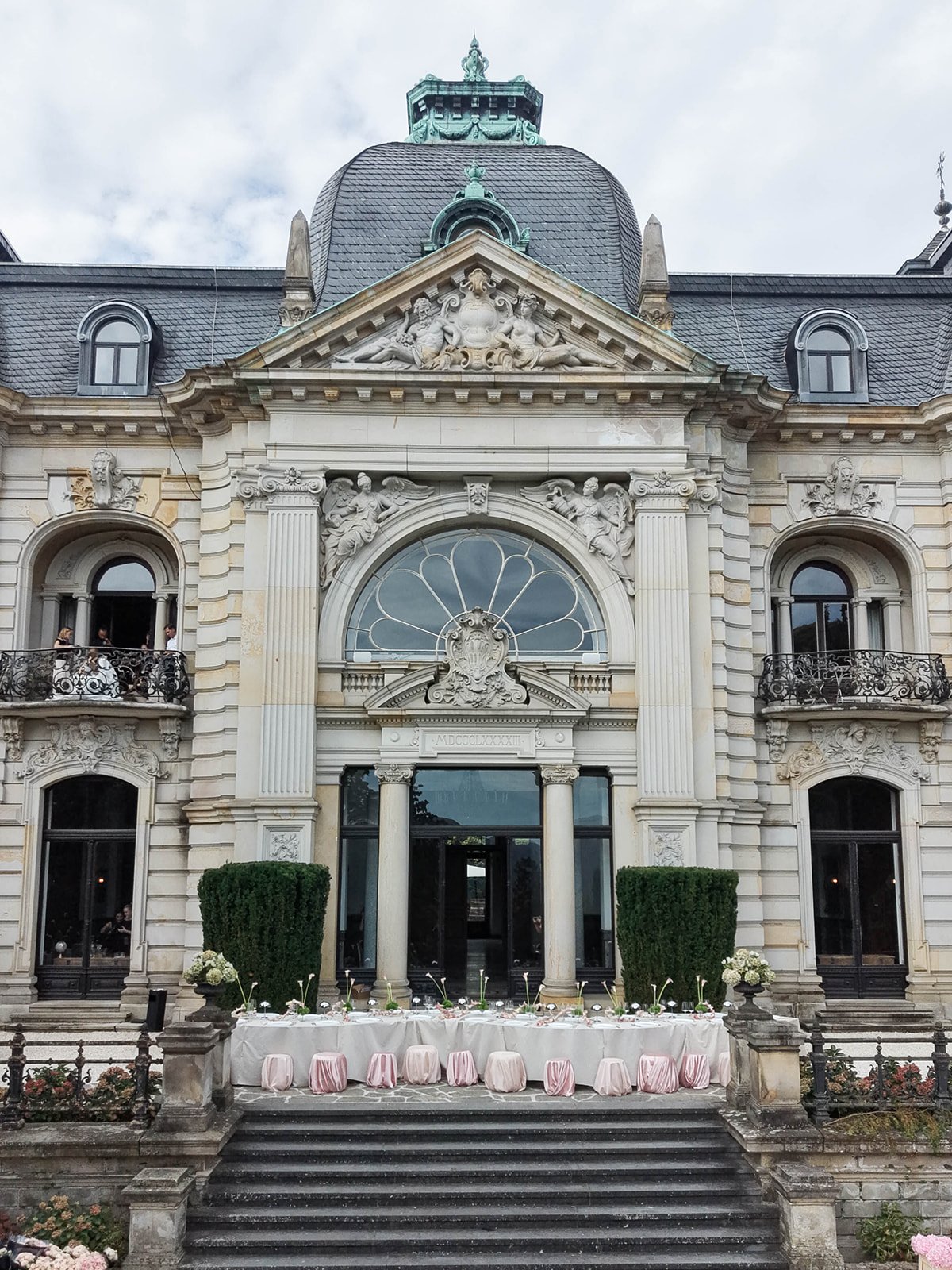 Front view of an ornate historic building with classical architecture, decorative sculptures, and large arched windows. There are two green bushes and a decorated table with pink covers and floral arrangements in front of the entrance. People are vis