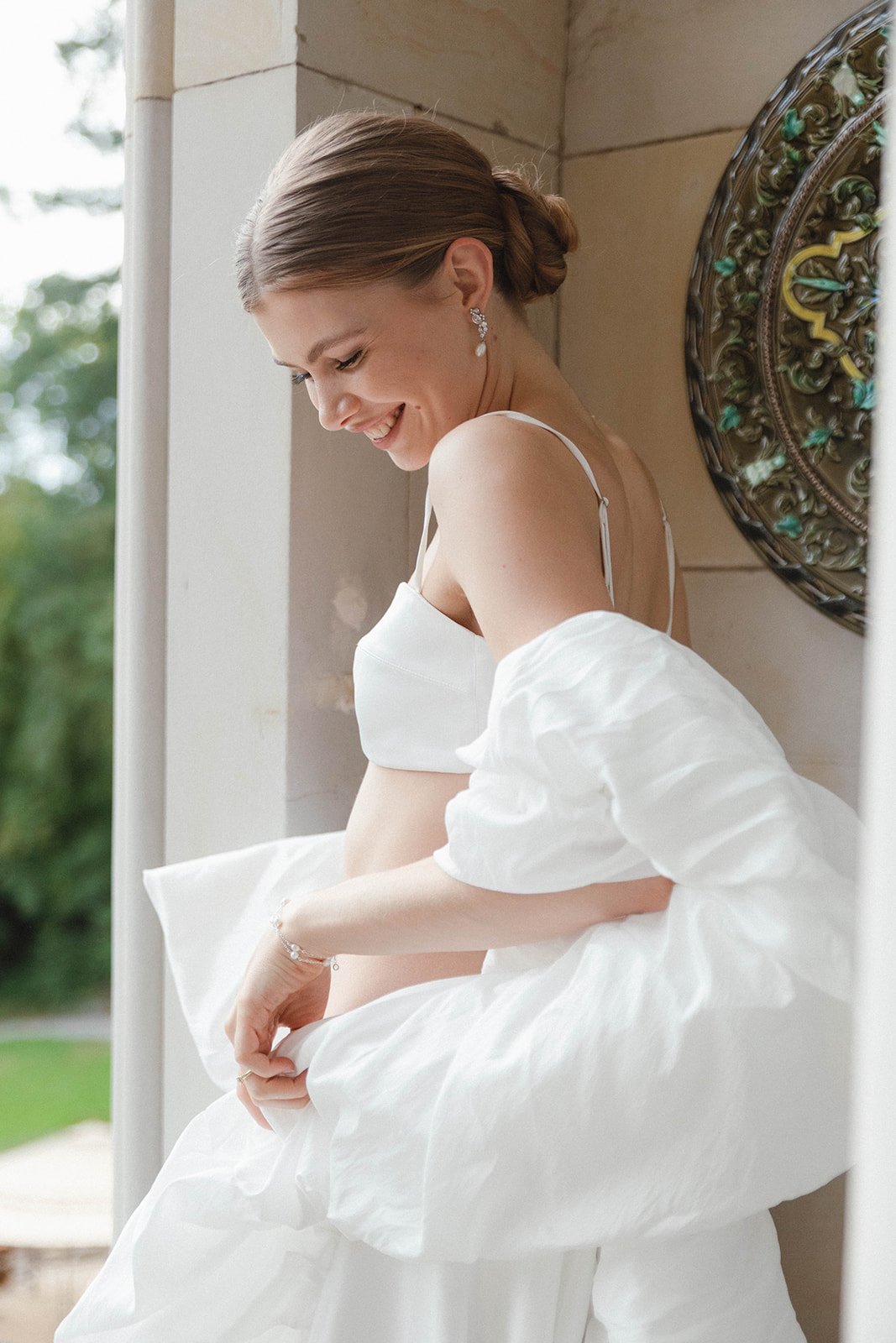 A woman with brown hair styled in an elegant bun, earrings, and a white dress, smiling and looking down while sitting on a ledge. Loose editorial style wedding photography.