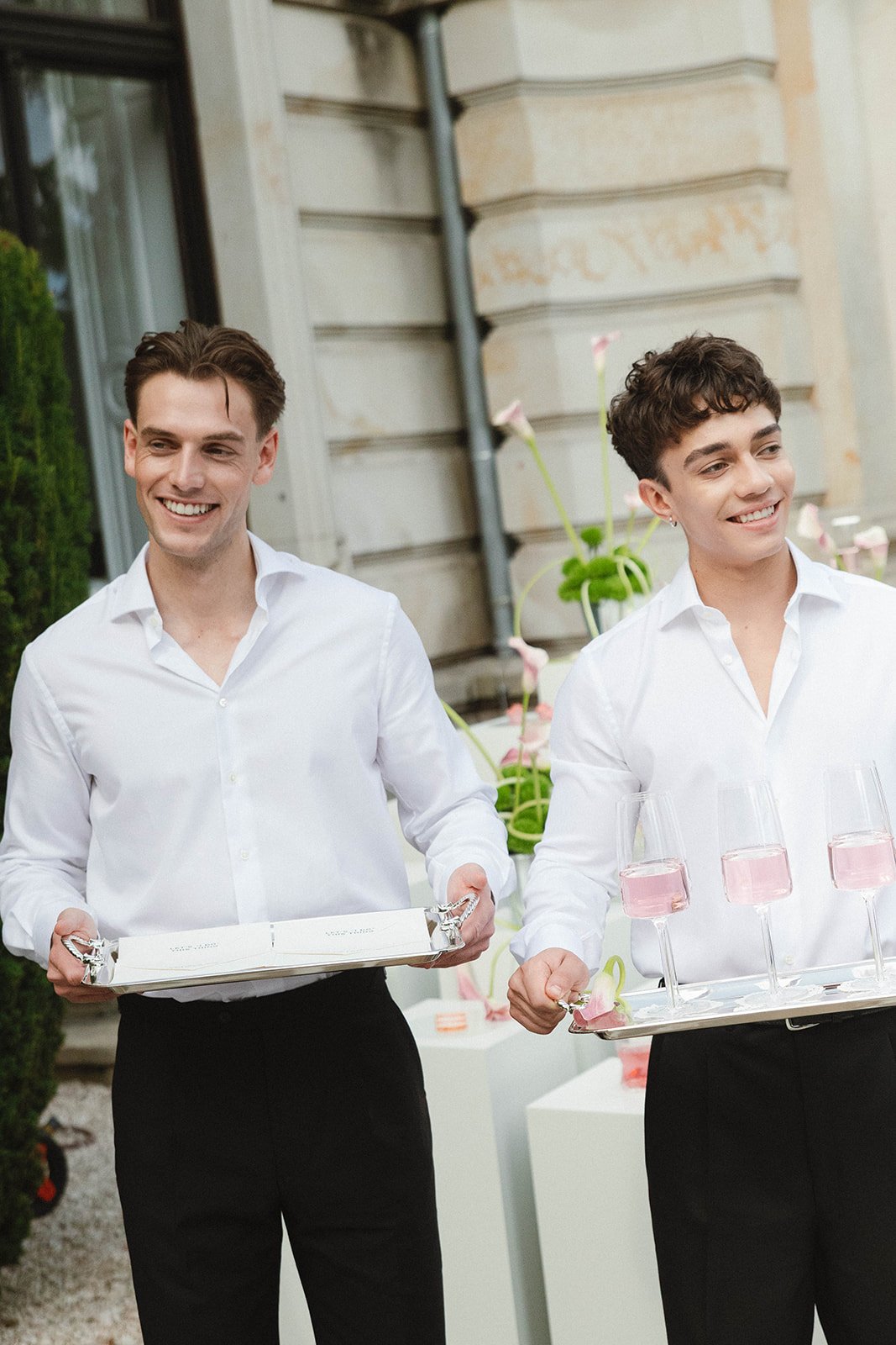 Two smiling male servers dressed in white shirts and black pants holding trays with glasses of pink drinks at an outdoor event.
