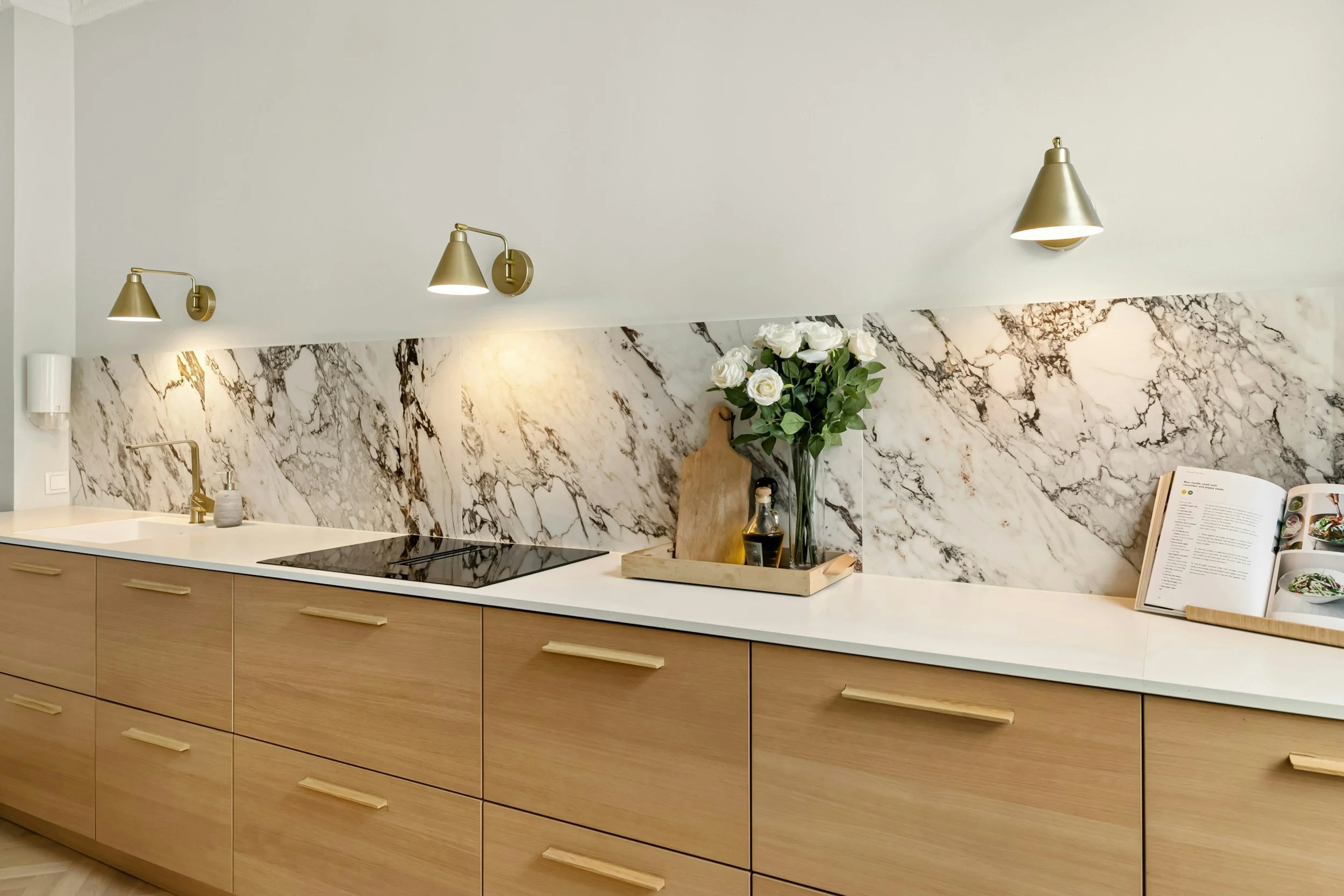 Modern kitchen with wooden cabinets, white marble backsplash, gold wall lamps, a countertop with a sink and electric stoves, a tray with a bouquet of white roses, a cutting board, a small jar, a bottle of oil, and an open cookbook.