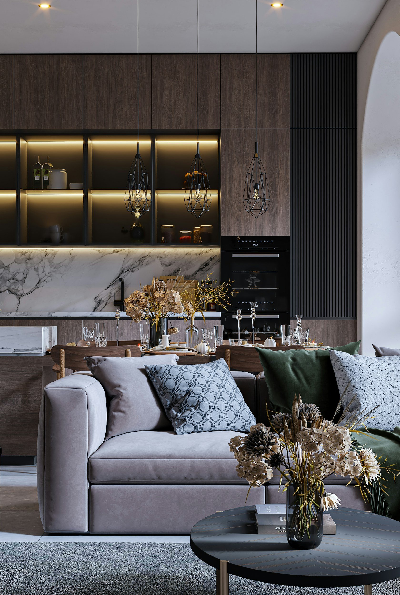 Modern living room with a beige sofa, decorative pillows, a coffee table with a vase of flowers, and a dining area in the background with a marble countertop, wooden cabinetry, pendant lights, and table settings.