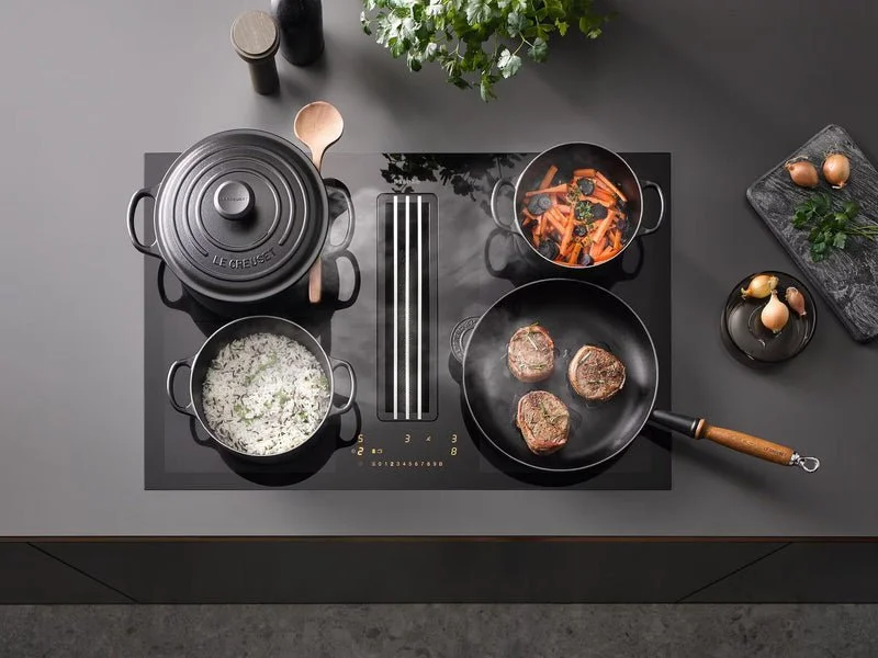 A modern stovetop with four pots and pans, one containing rice, another with carrots and spinach, a third with cooking steaks, and a lid on a small pot. There are eggs and green herbs on a black cutting board, and a plant in the background.