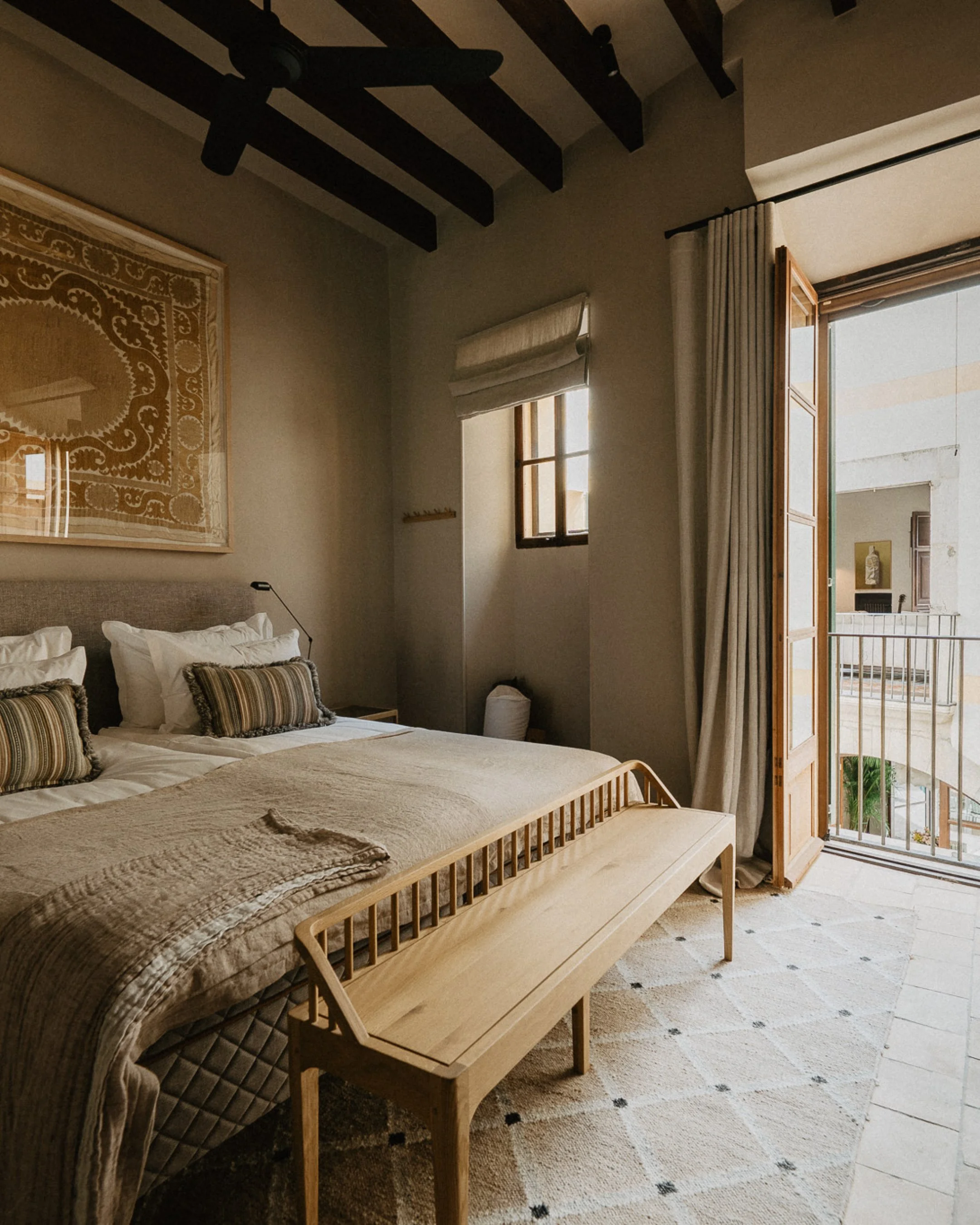 A bedroom with a large bed, beige bedding, striped pillows, wooden bench at the foot, and a balcony door open to a small outdoor space with a metal railing and neighboring buildings.
