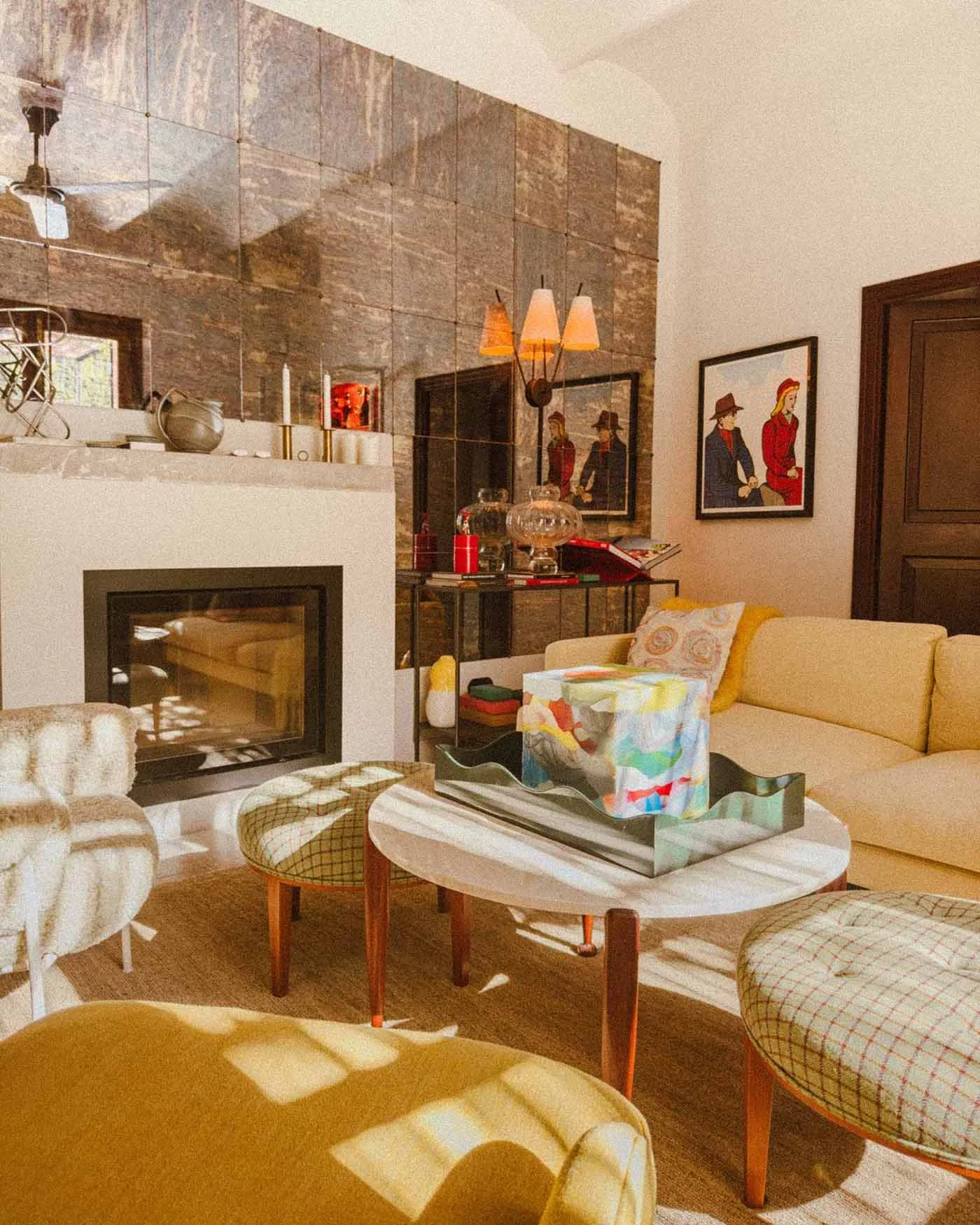Cozy living room with a yellow sofa, patterned chairs, a white coffee table with a colorful box, and a modern fireplace. Decor includes framed art, a table with glass lamps, and wall-mounted lighting, with a stone accent wall and a window letting in natural light.