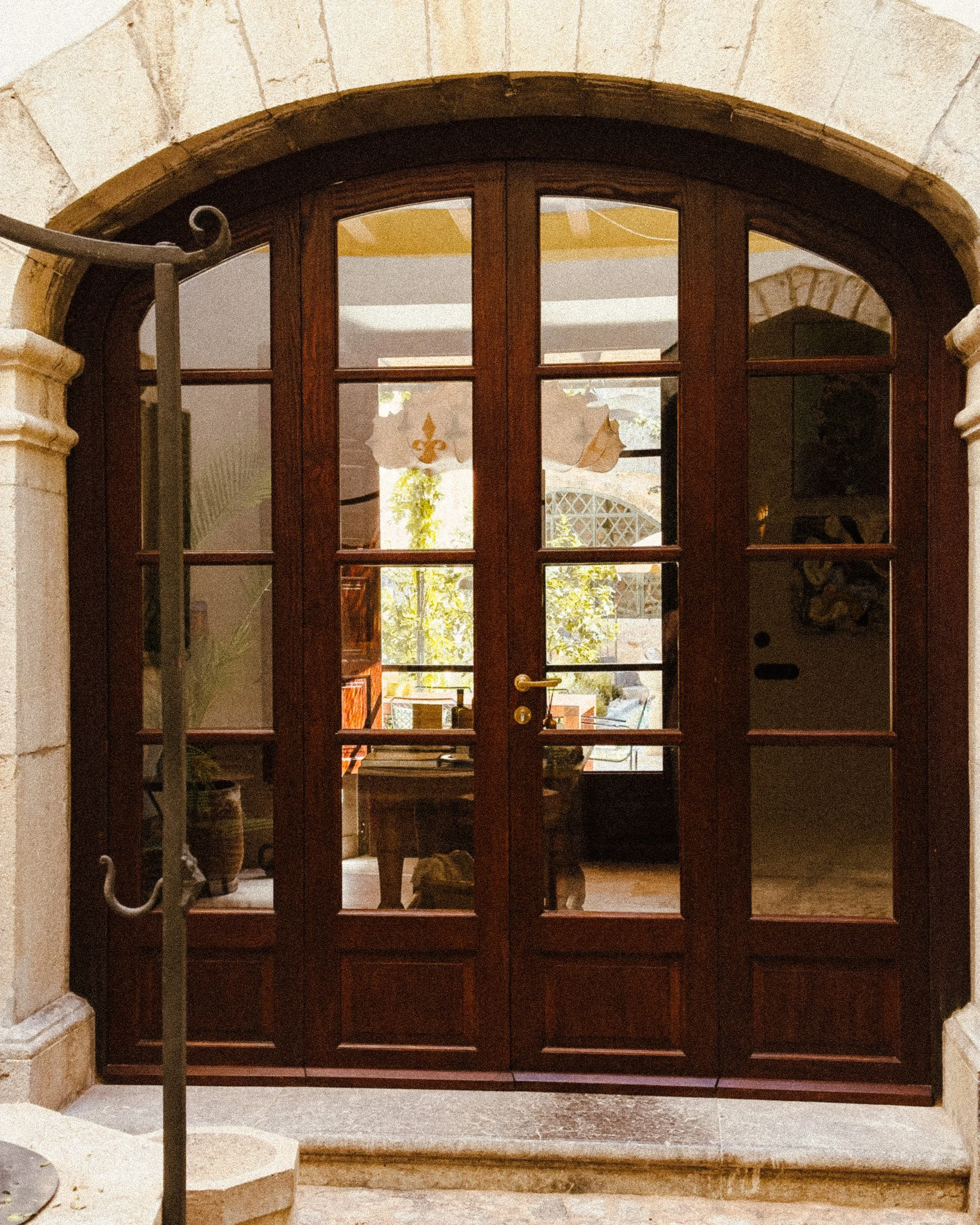 Wooden-framed glass double doors leading to an interior room with a table, decorative hanging banner, potted plant, and artwork, viewed from an arched stone doorway.