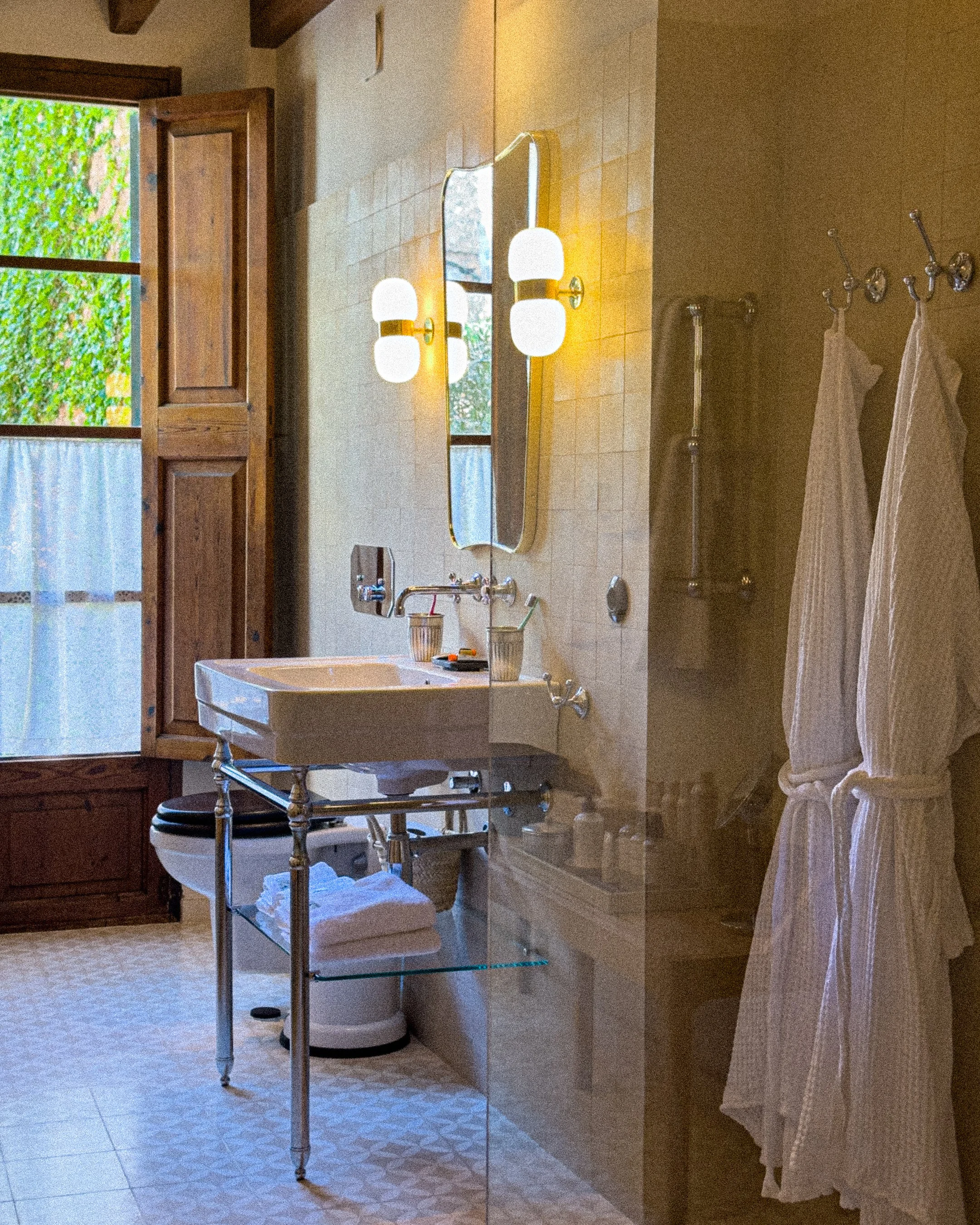 A vintage bathroom with a window, open wooden shutters, a wall-mounted mirror with white globe lights, a white sink with chrome fixtures, a glass shelf with towels, and two frosted glass robes hanging on hooks.