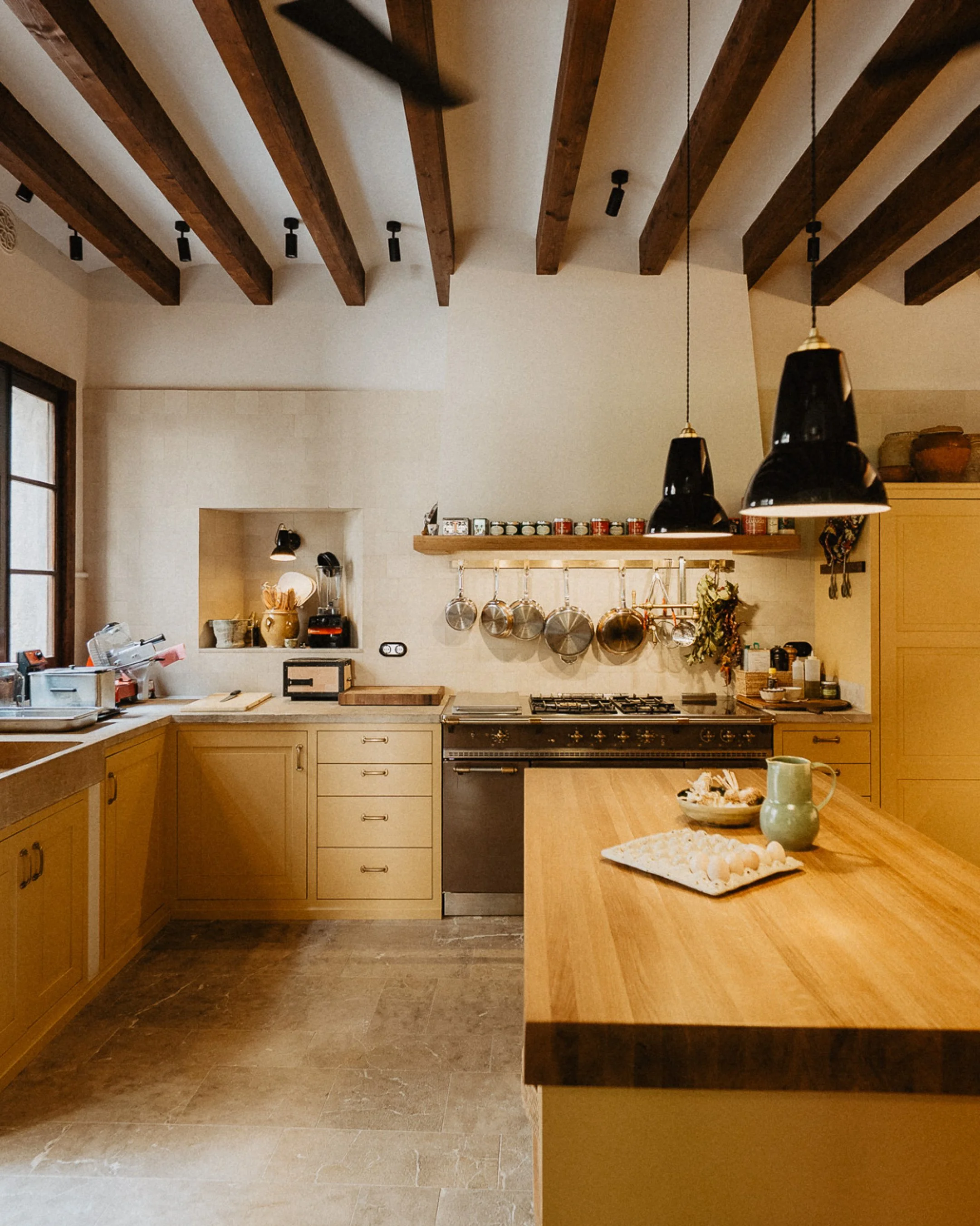 A kitchen with wooden cabinets, hanging pots, black pendant lights, and a wooden countertop with a green pitcher and bowl of eggs.