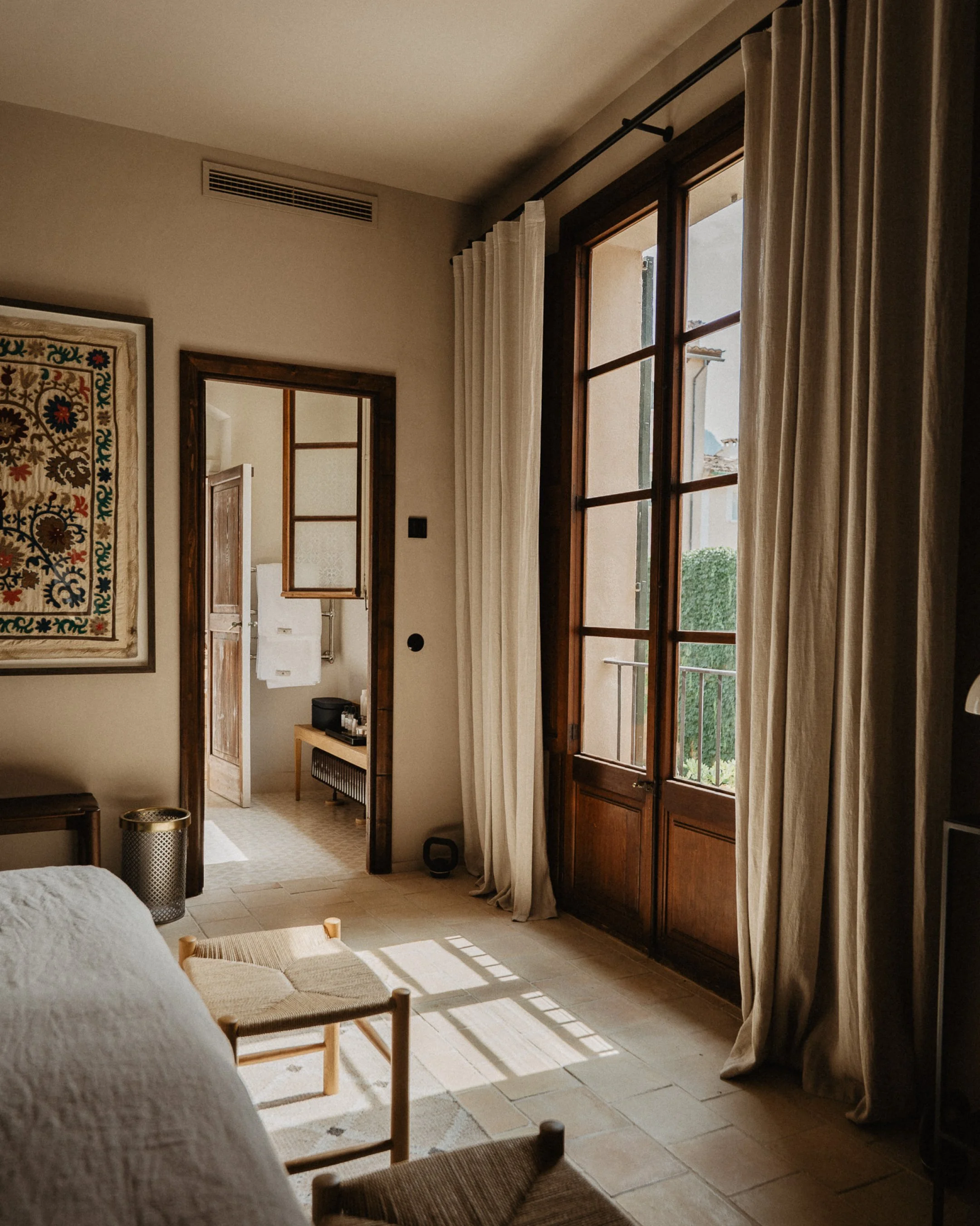 A cozy bedroom with a door leading to a balcony, large windows with white curtains, a bed, a bench, and a view of a garden outside.