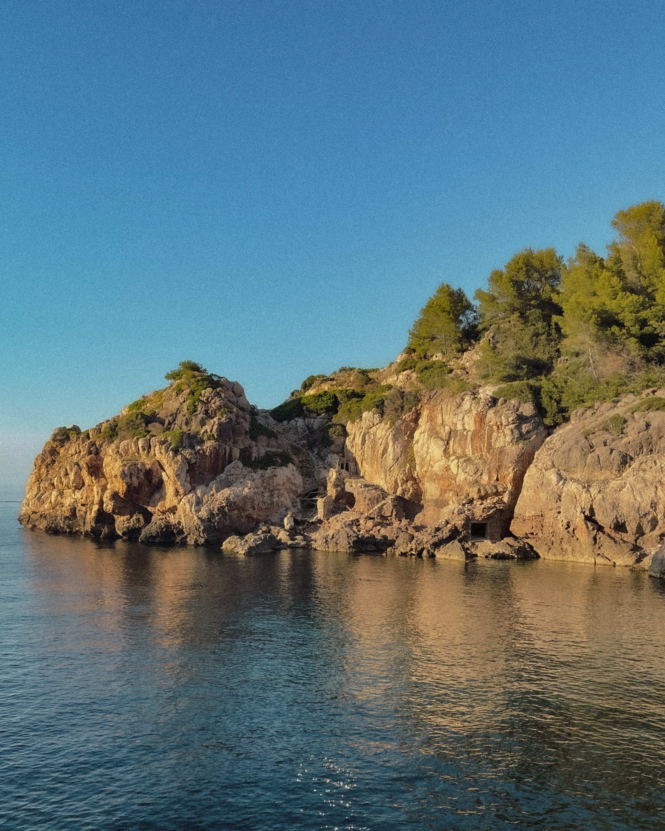 A rocky coastline with green trees on top, calm water in the foreground, and a clear blue sky above.