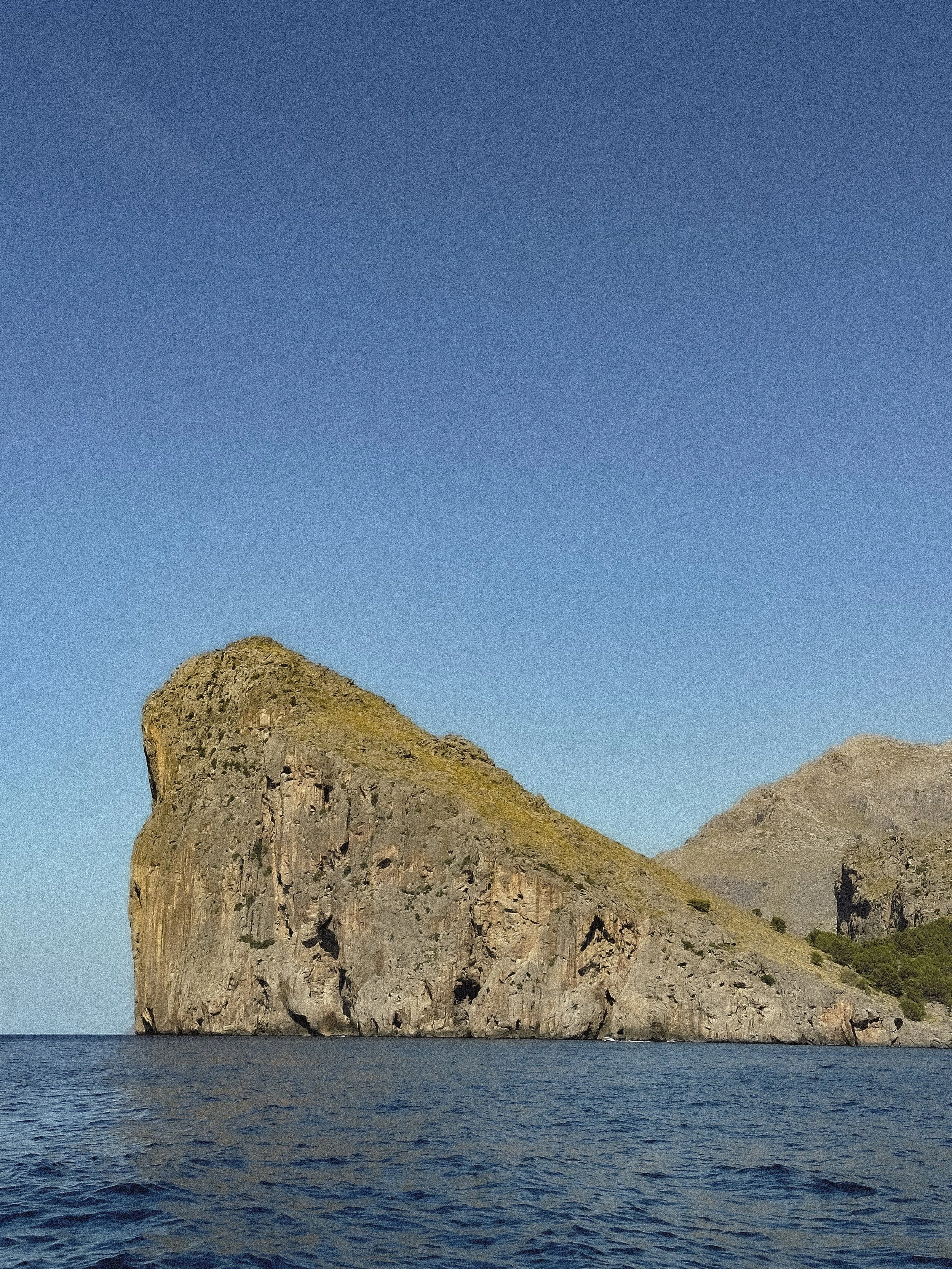 A large rocky island with steep cliffs rising from a calm ocean, under a clear blue sky.