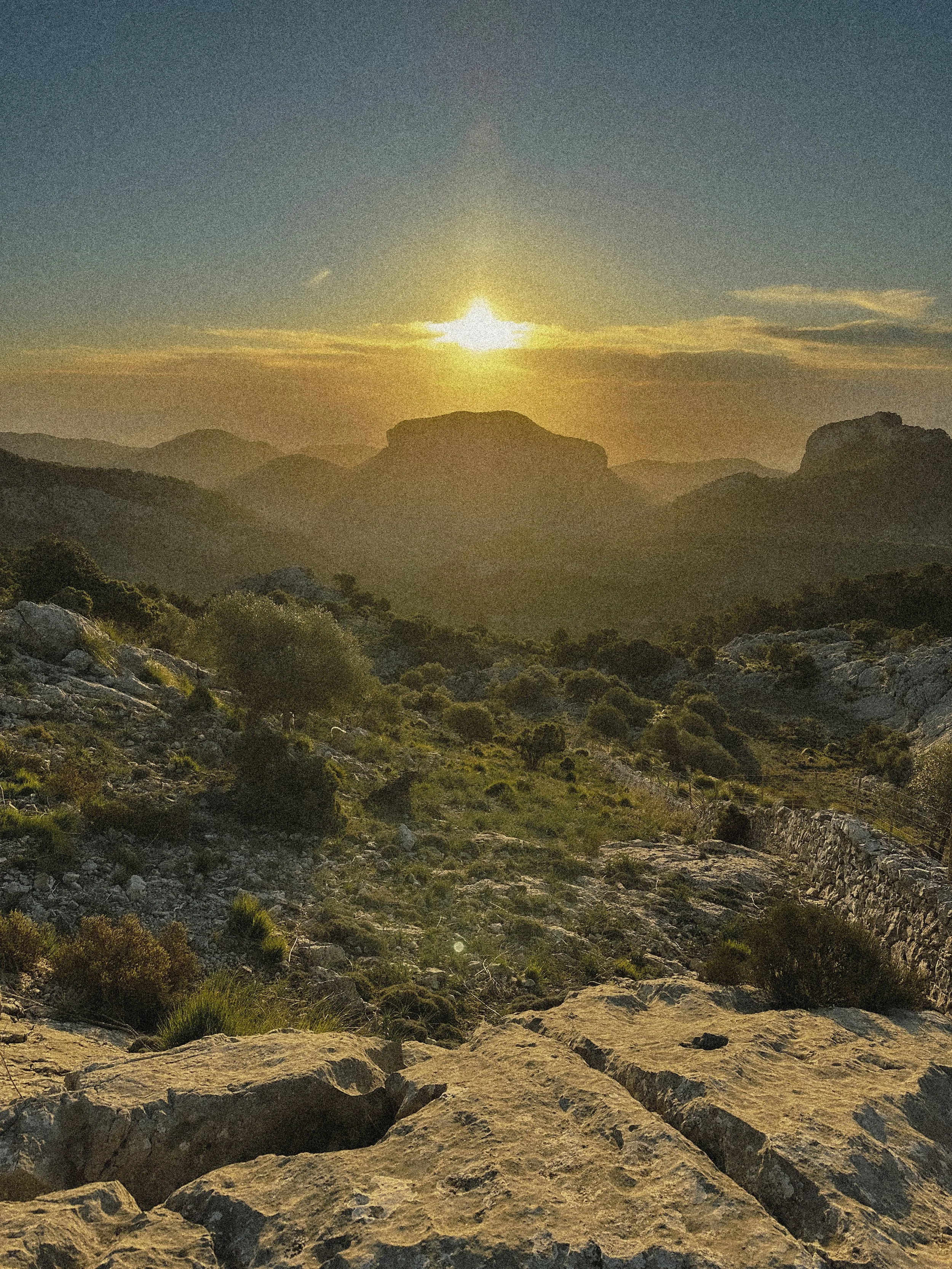 Sunset over a mountainous landscape with rocky terrain and scattered bushes.