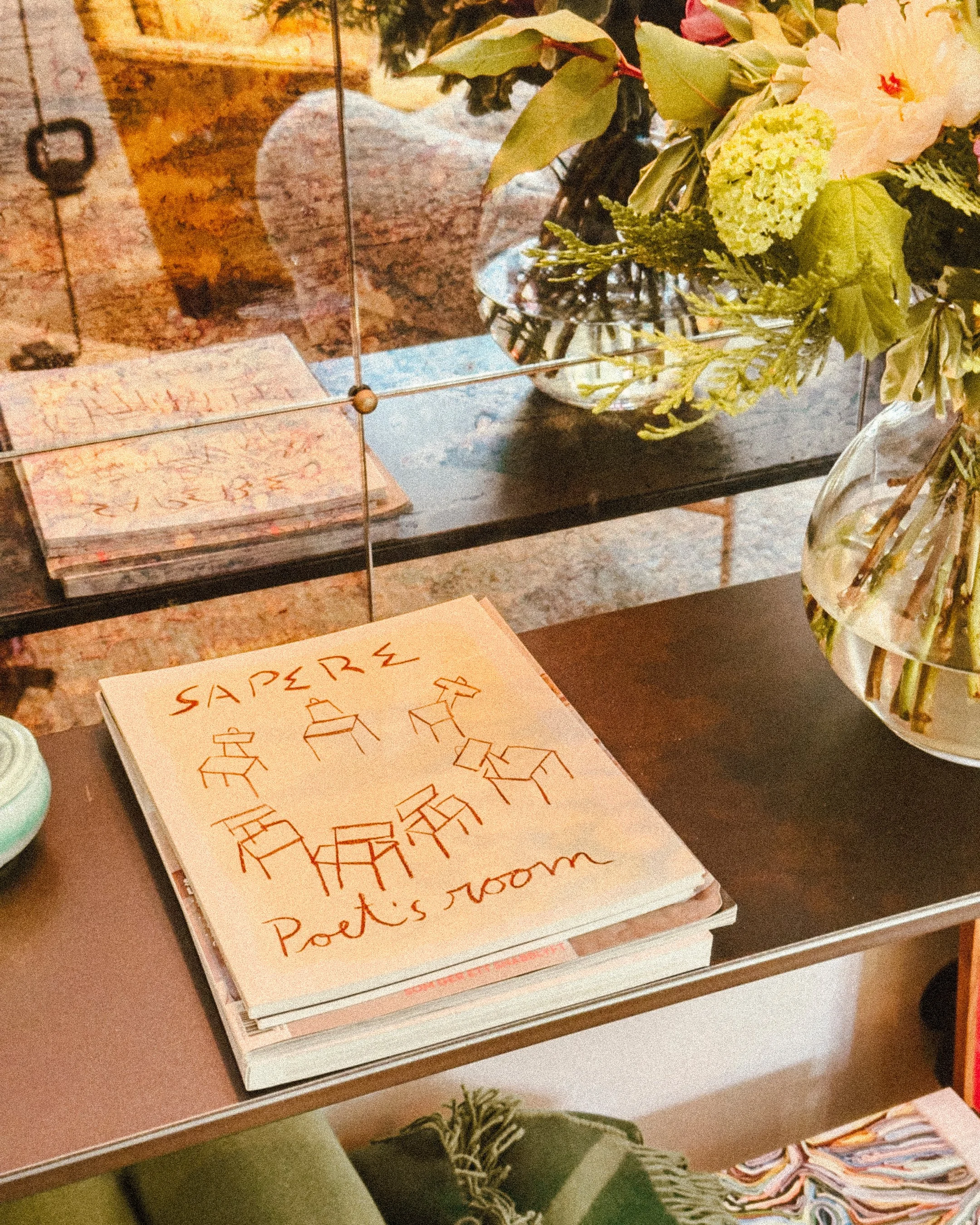 A notebook with the words 'SAPPERS' and 'Poet's room' written on the cover, along with simple line drawings of chairs, a table, and a lamp, placed on a shelf next to a vase of flowers.