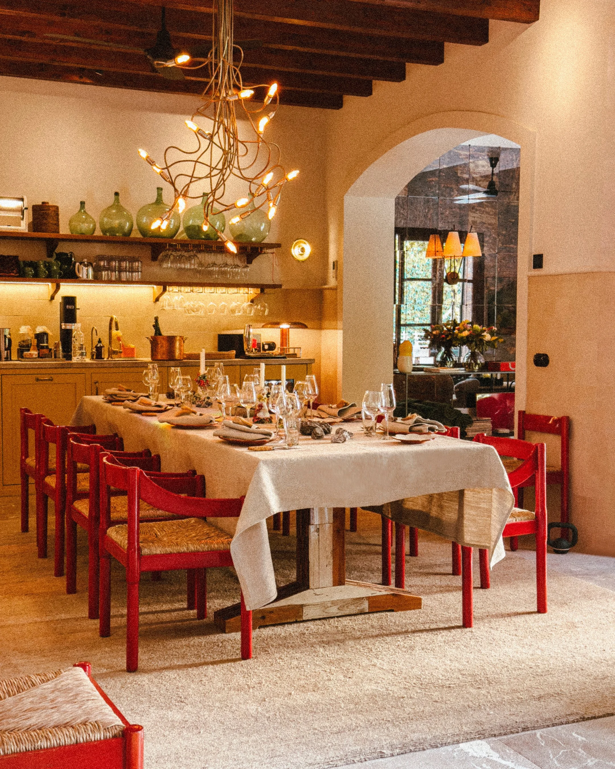 A dining room with a long table set for a meal, decorated with wine glasses, plates, and napkins, surrounded by red chairs. The room has a wooden ceiling with a modern chandelier, a kitchen area with shelves, green vases, and glassware, and a window with a vase of flowers nearby.