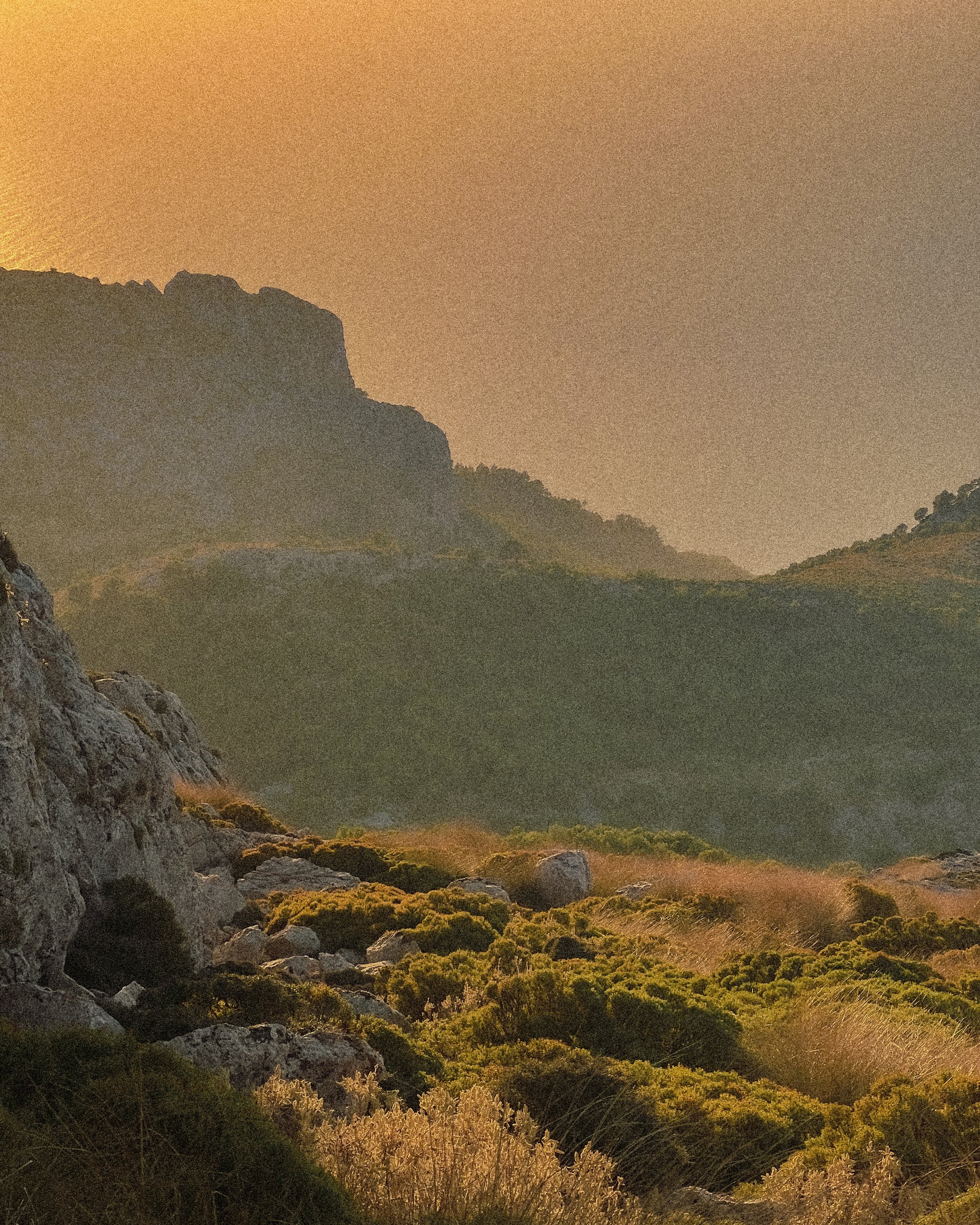 Mountain landscape during sunset with rocky foreground, green shrubs, and rolling hills under an orange sky.