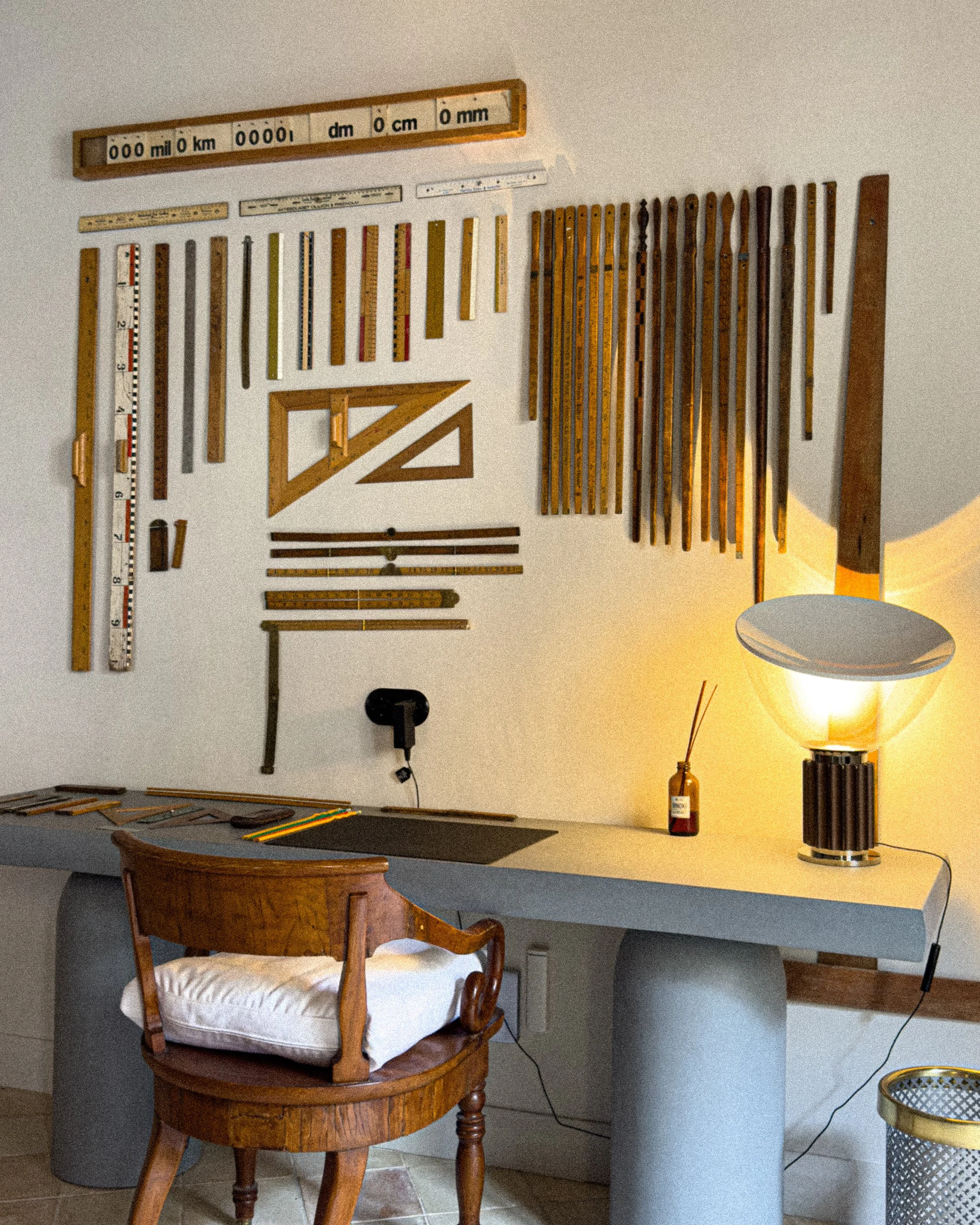 A workspace with carpentry tools and measures on a wall, a vintage wooden chair with a cushion, a gray desk, a lamp, and a small bottle on the desk.