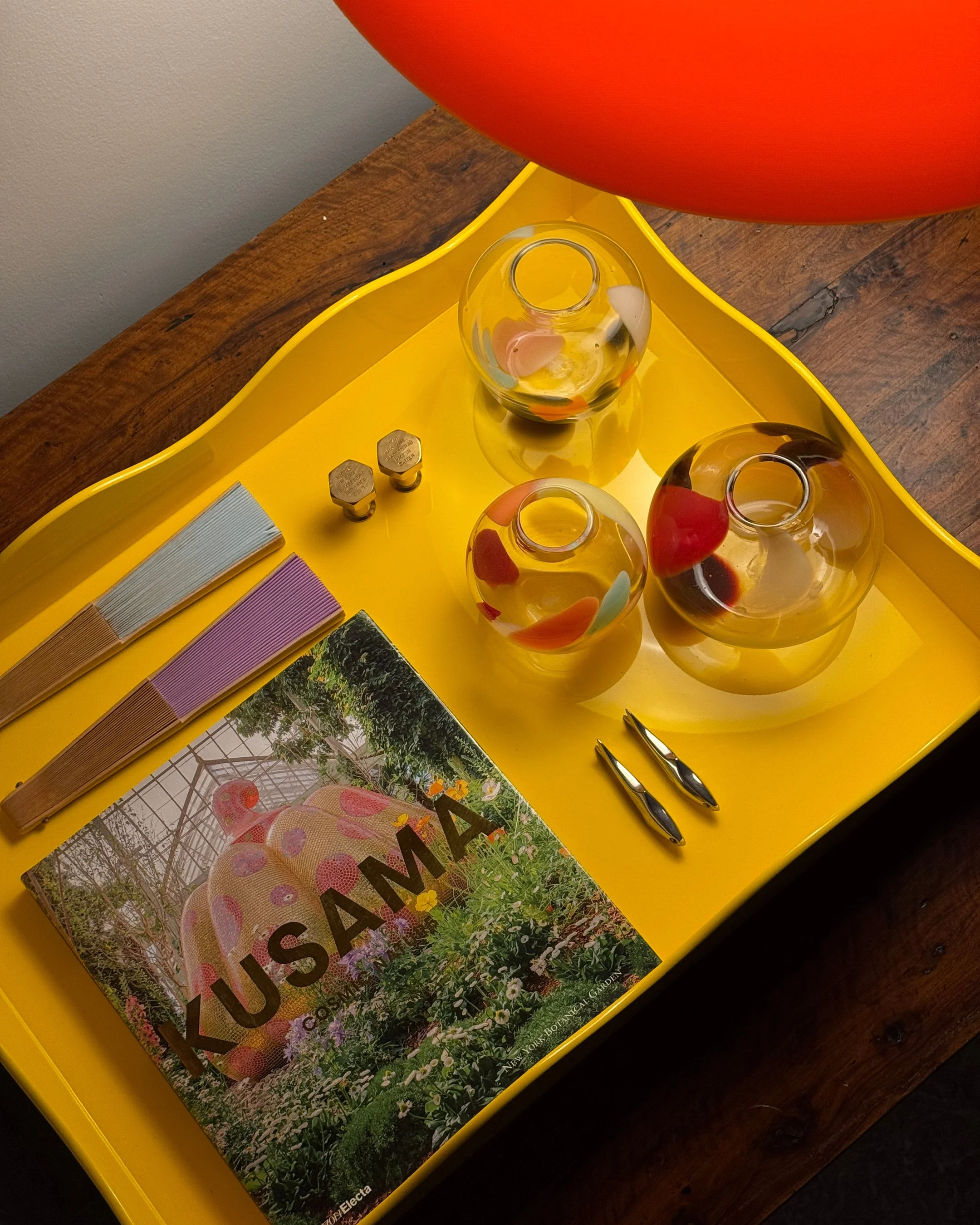 Yellow tray on a wooden table holding three glass vases with colorful abstract designs, a KUSAMA book with floral cover art, two small metal objects, two metallic pens, and some combs or hair clips.