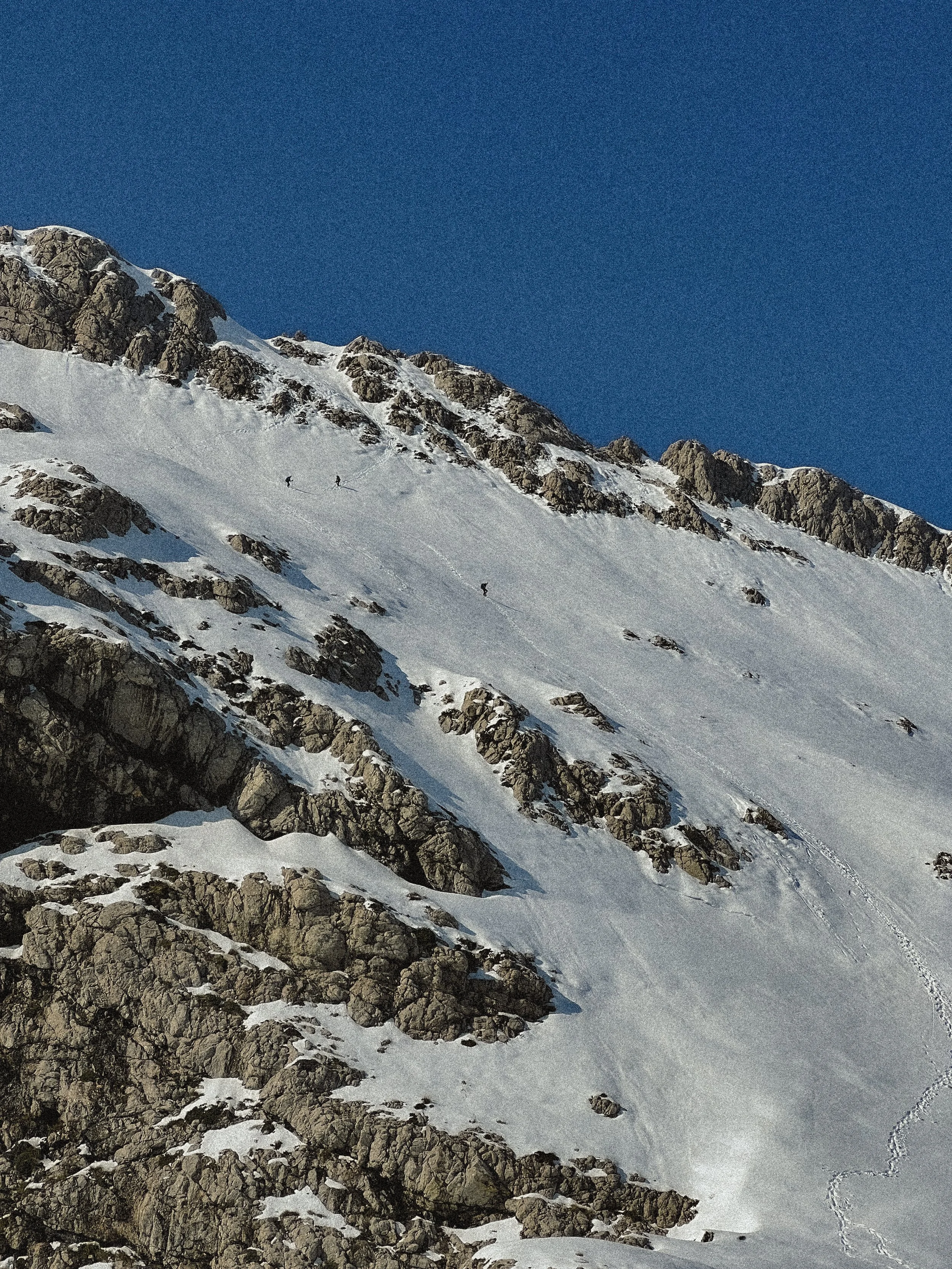 Snow-covered mountain slope with rocky outcroppings and a few small climbers ascending.